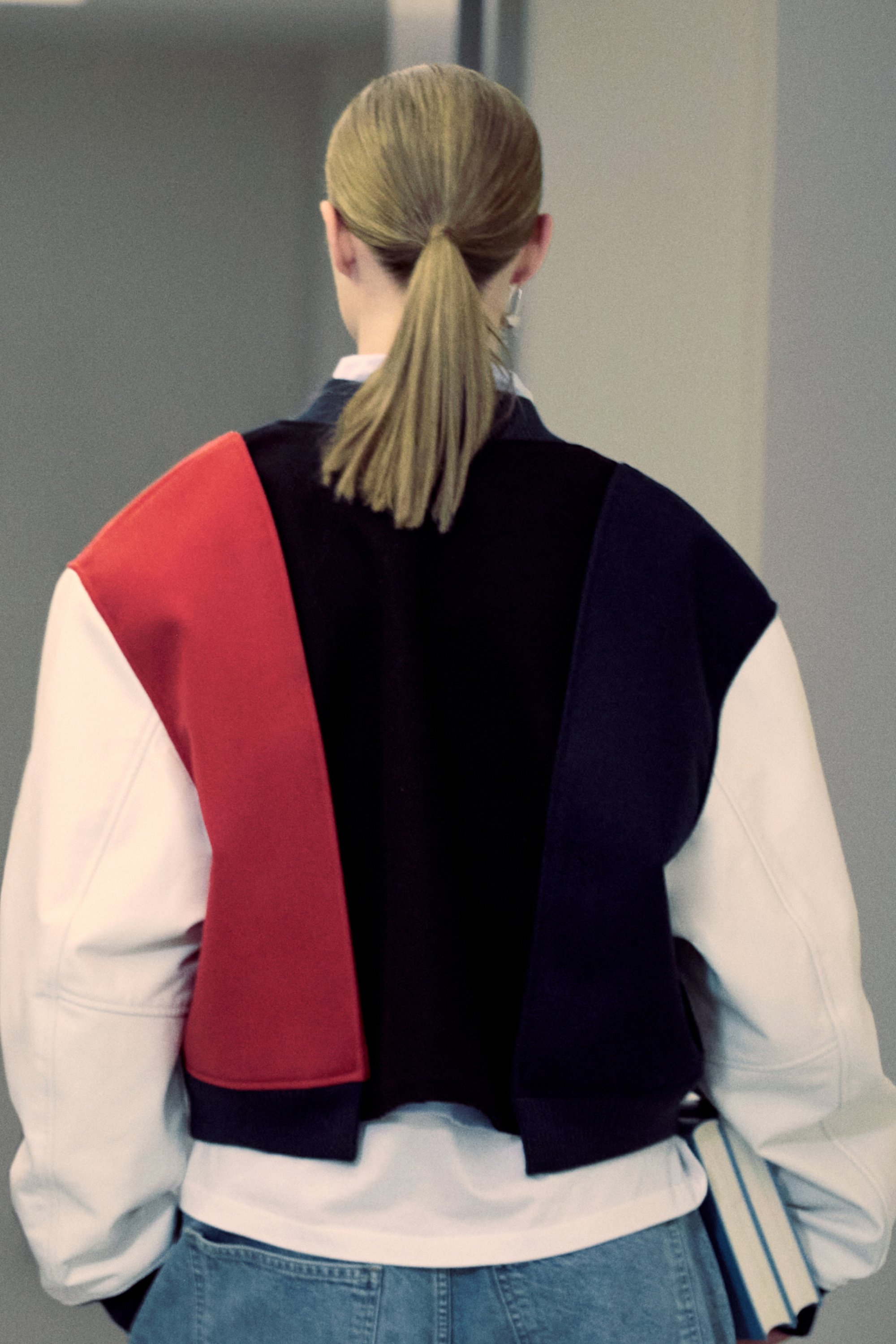Una persona con lunghi capelli biondi raccolti in una coda di cavallo indossa una grande giacca color block con sezioni rosse, nere e bianche, mentre tiene dei libri.
