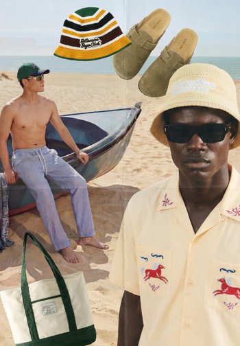 Homme torse nu en pantalon rayé près d'un bateau sur la plage, homme à la peau foncée portant une chemise brodée, chapeaux bob, chaussures en daim sans lacets et sac en toile.