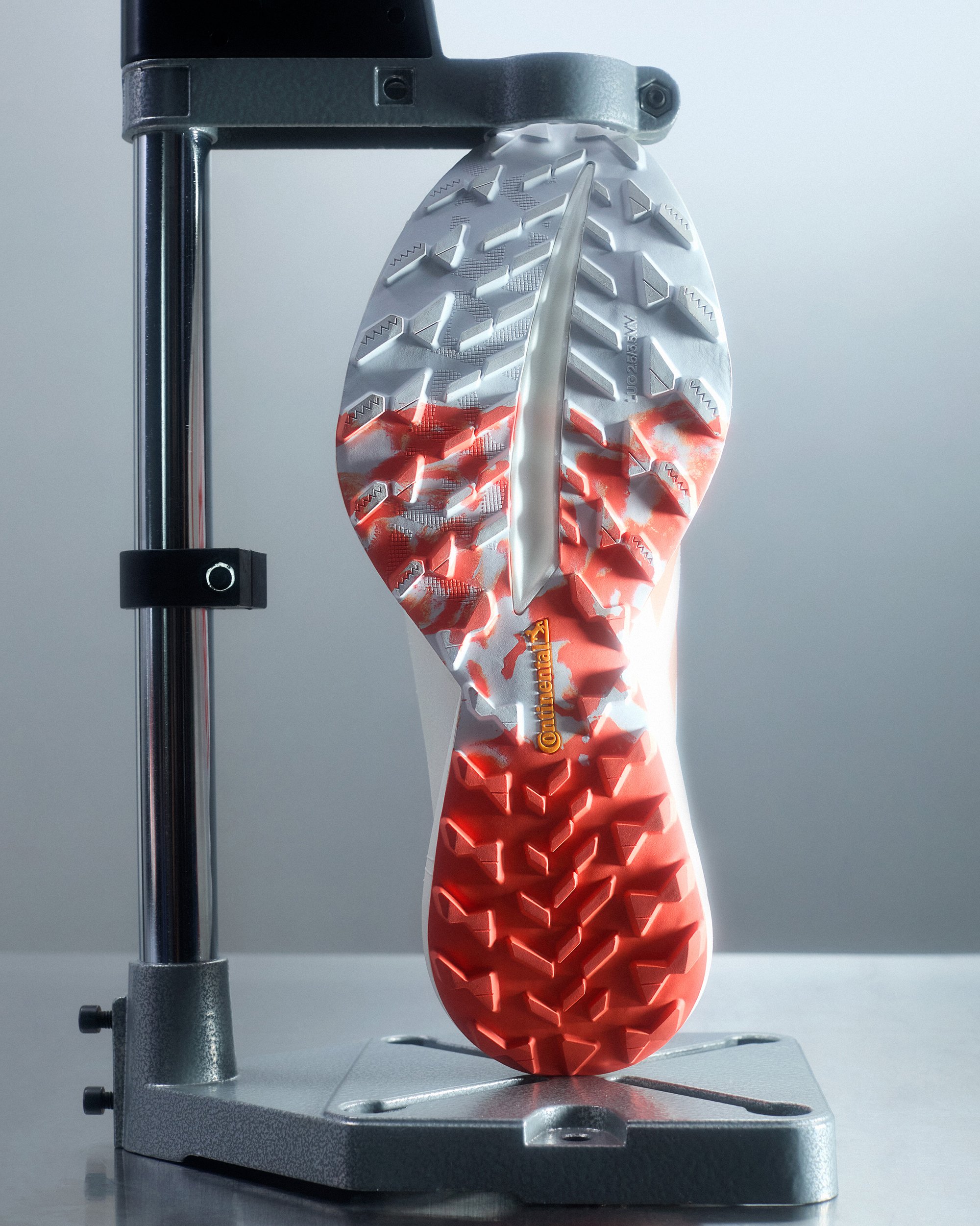 Sole of a running shoe displayed upright, showcasing intricate tread patterns in red and white, mounted on a metallic fixture.