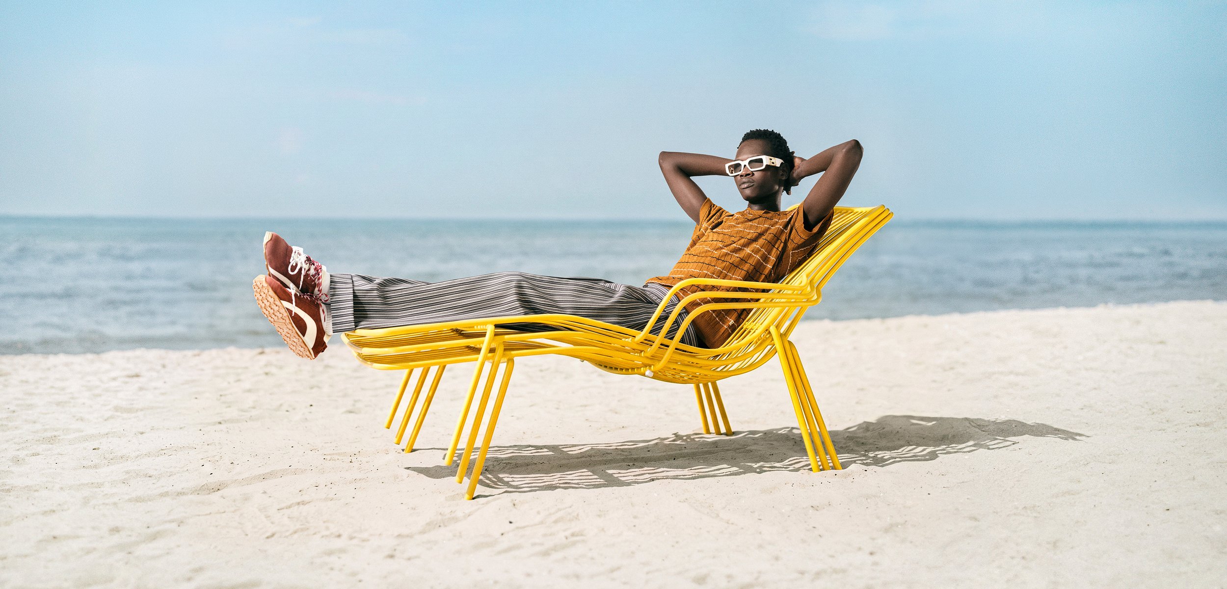 Person mit Sonnenbrille und Freizeitkleidung entspannt sich auf einem gelben Liegestuhl an einem Sandstrand mit ruhigem Meer im Hintergrund.