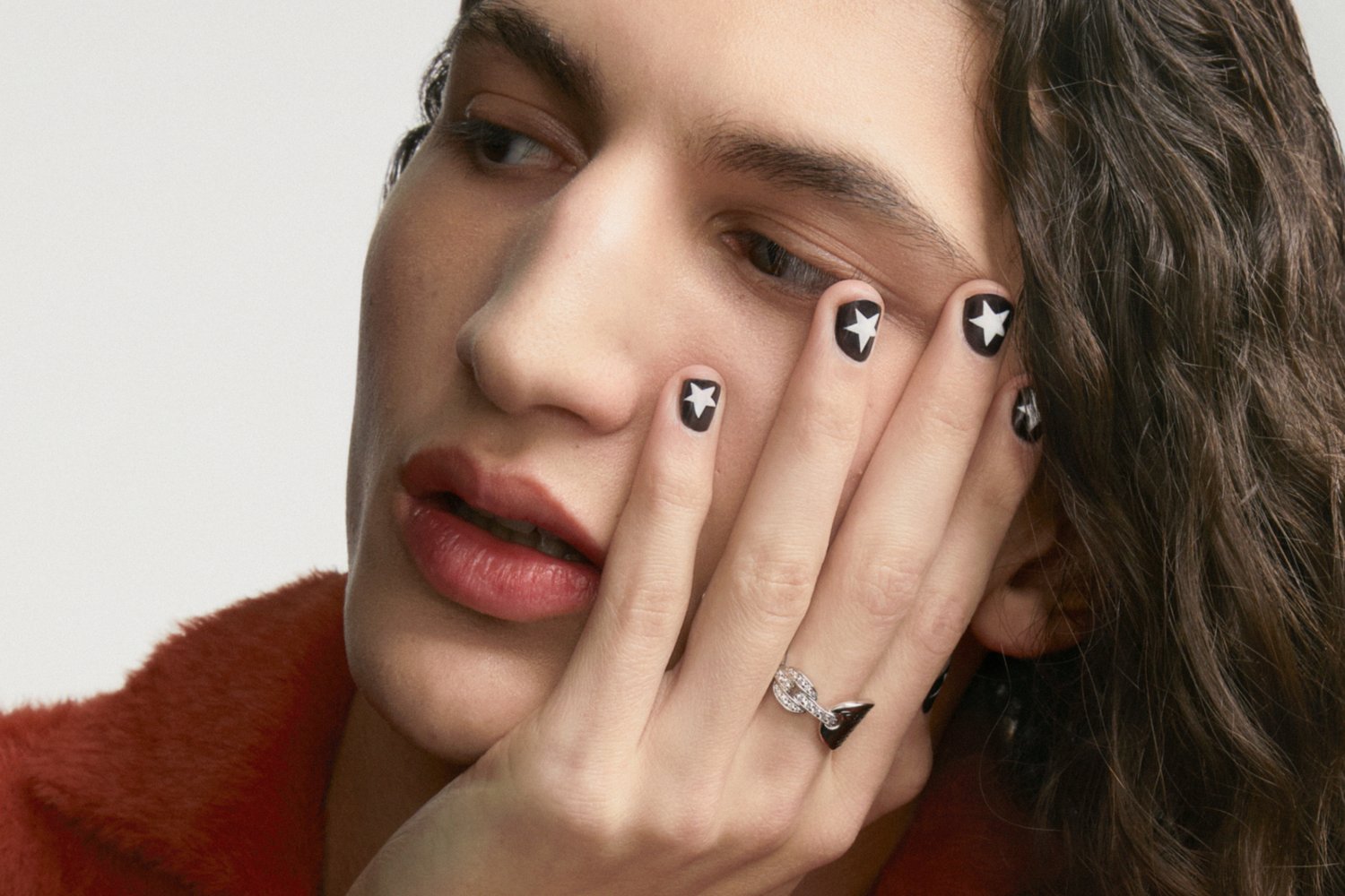 A person with wavy hair rests their face on one hand, showcasing black star-patterned nails and a silver ring on the opposite hand.