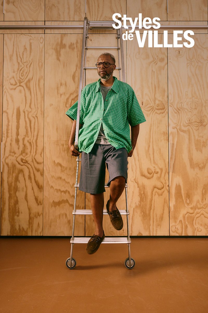 Homme portant des lunettes et une chemise verte à carreaux assis sur une échelle en métal contre un mur en panneaux de bois avec le texte "Styles de Villes".