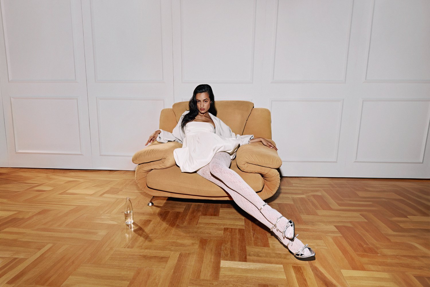 Una persona con un vestido blanco se relaja en un sofá beige, vistiendo unas medias con estampado y zapatos elegantes, con una botella de vidrio en primer plano.