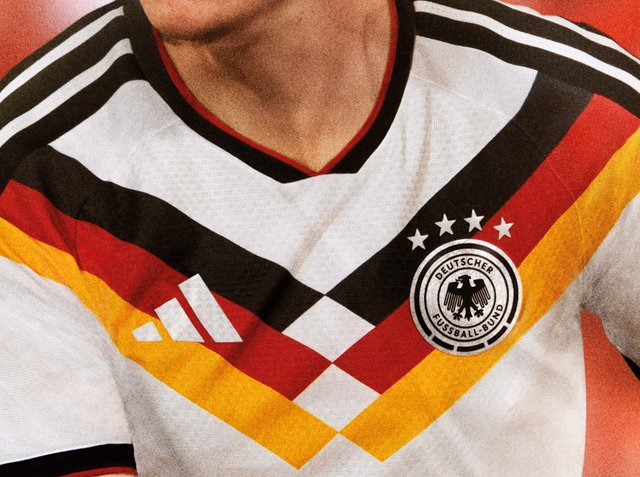 Zbliżenie na osobę w białej koszulce piłkarskiej reprezentacji Niemiec z czarnymi, czerwonymi i żółtymi paskami oraz logo Deutscher Fussball-Bund.