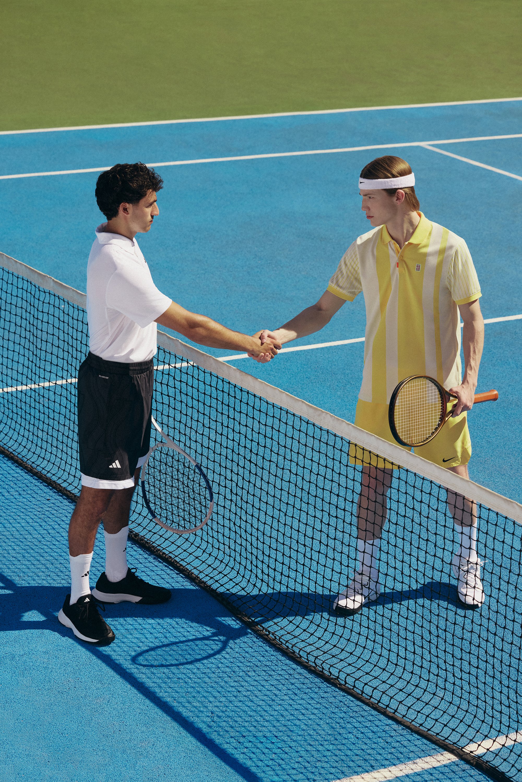 Dos jóvenes se dan la mano a través de una red en una cancha de tenis, uno lleva una camiseta blanca con pantalones cortos negros, el otro un conjunto a rayas amarillas.