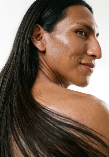 Profil af en person med langt, glat sort hår, solbrun hud og et lille smil, vendt let over skulderen mod en hvid baggrund.