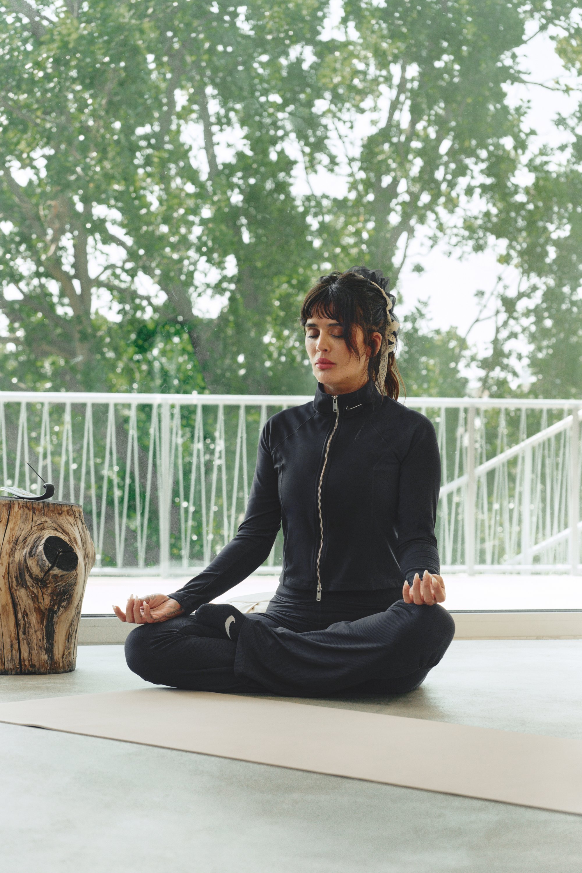 Chaqueta atlética negra con cremallera y pantalones a juego, sentada en una postura de piernas cruzadas sobre una esterilla de yoga, con luz natural y vegetación visible.