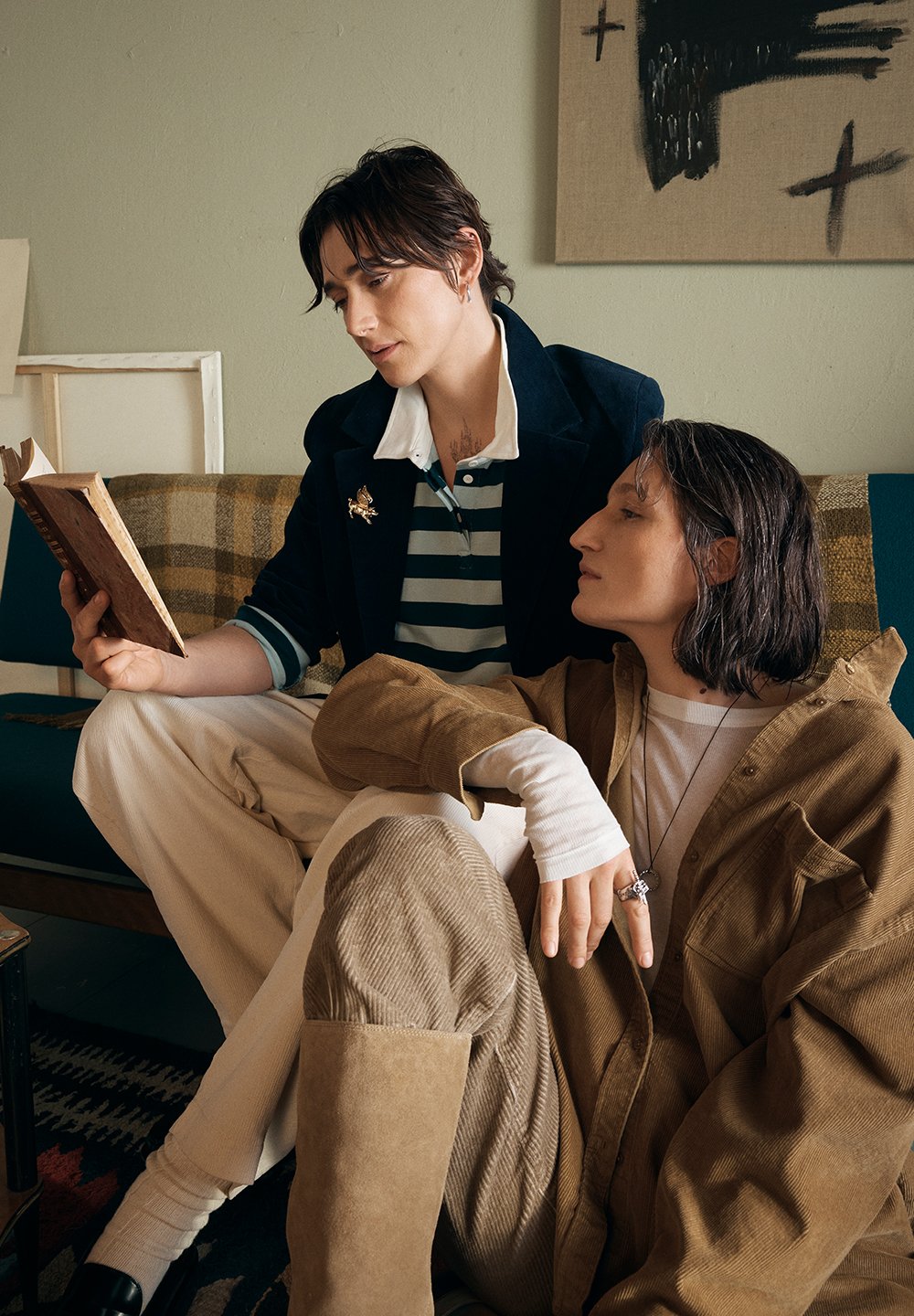 Két személy ül egy kanapén. Az egyik egy tengerészkék blézert visel fehér gallérral és csíkos inggel, és éppen egy könyvet olvas. A másik barna kordbársony zakóban van, világos inggel és egy kiegészítő gyűrűvel.