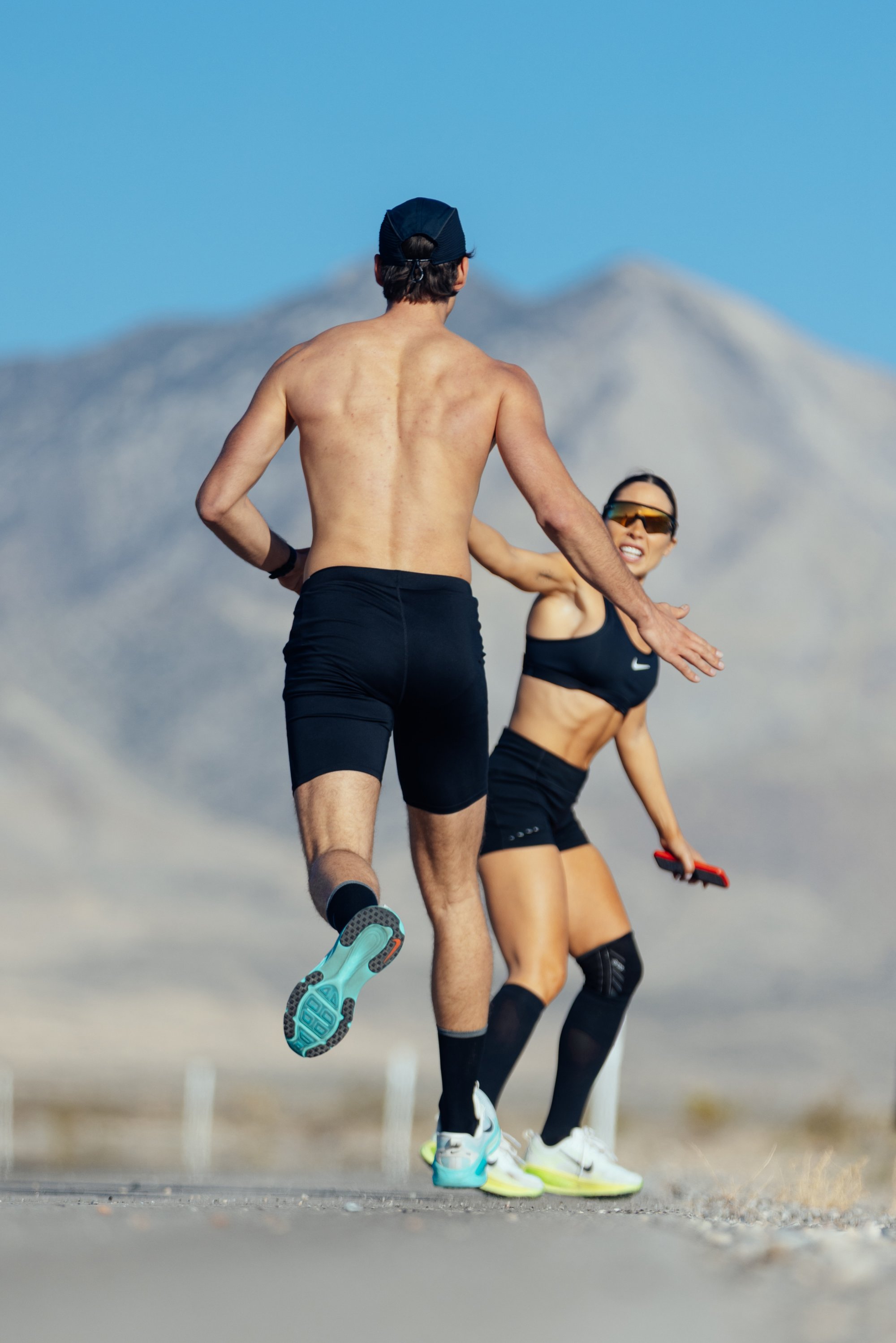 Two runners on a sunny road engage in a high-five. One is shirtless, while the other wears a sports top and shorts, smiling. Mountains loom behind.