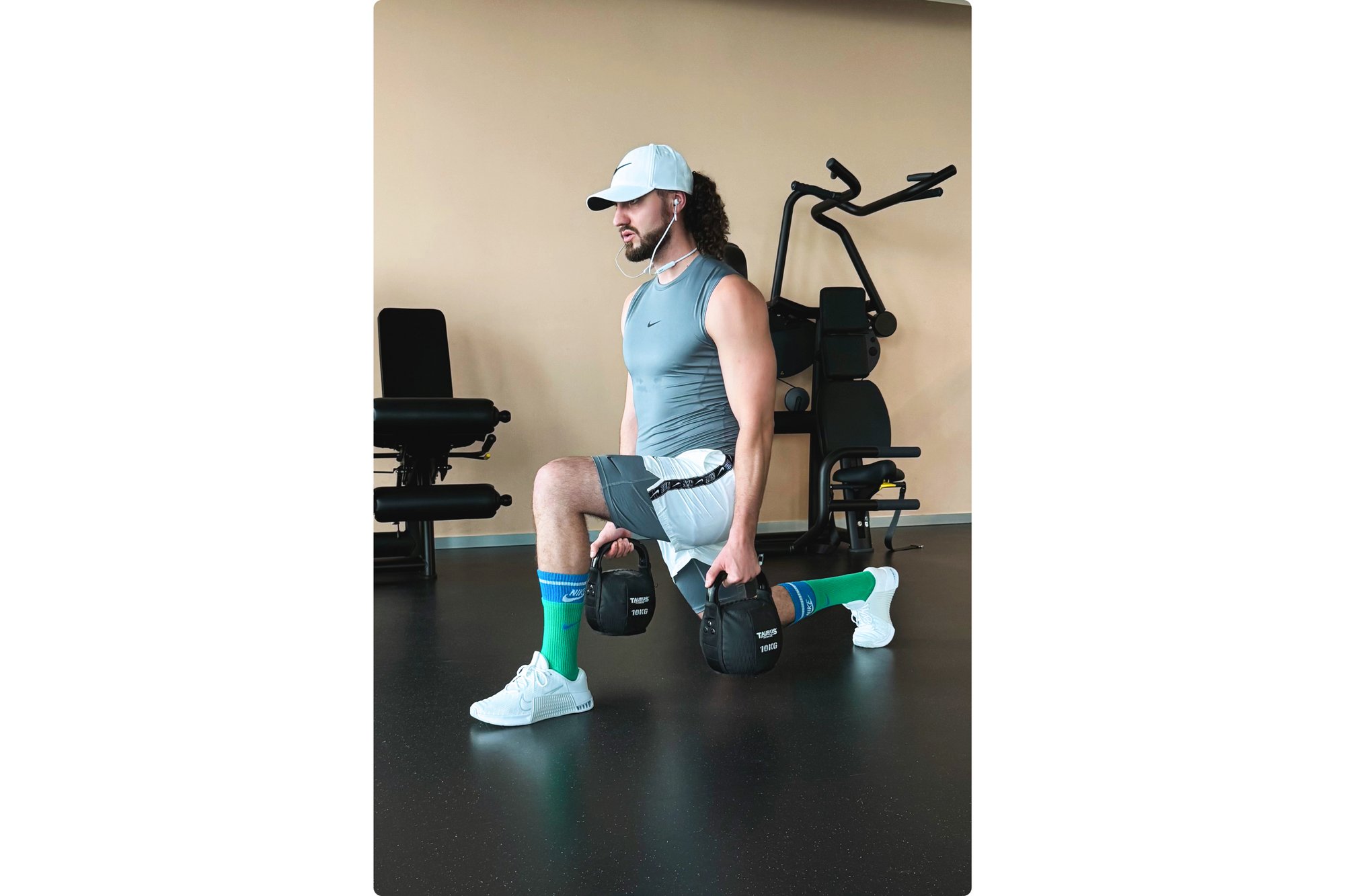 Un hombre realiza una zancada en un gimnasio, sosteniendo pesas pesadas, vistiendo una camiseta ajustada, pantalones cortos y calcetines coloridos, con equipo de entrenamiento en el fondo.