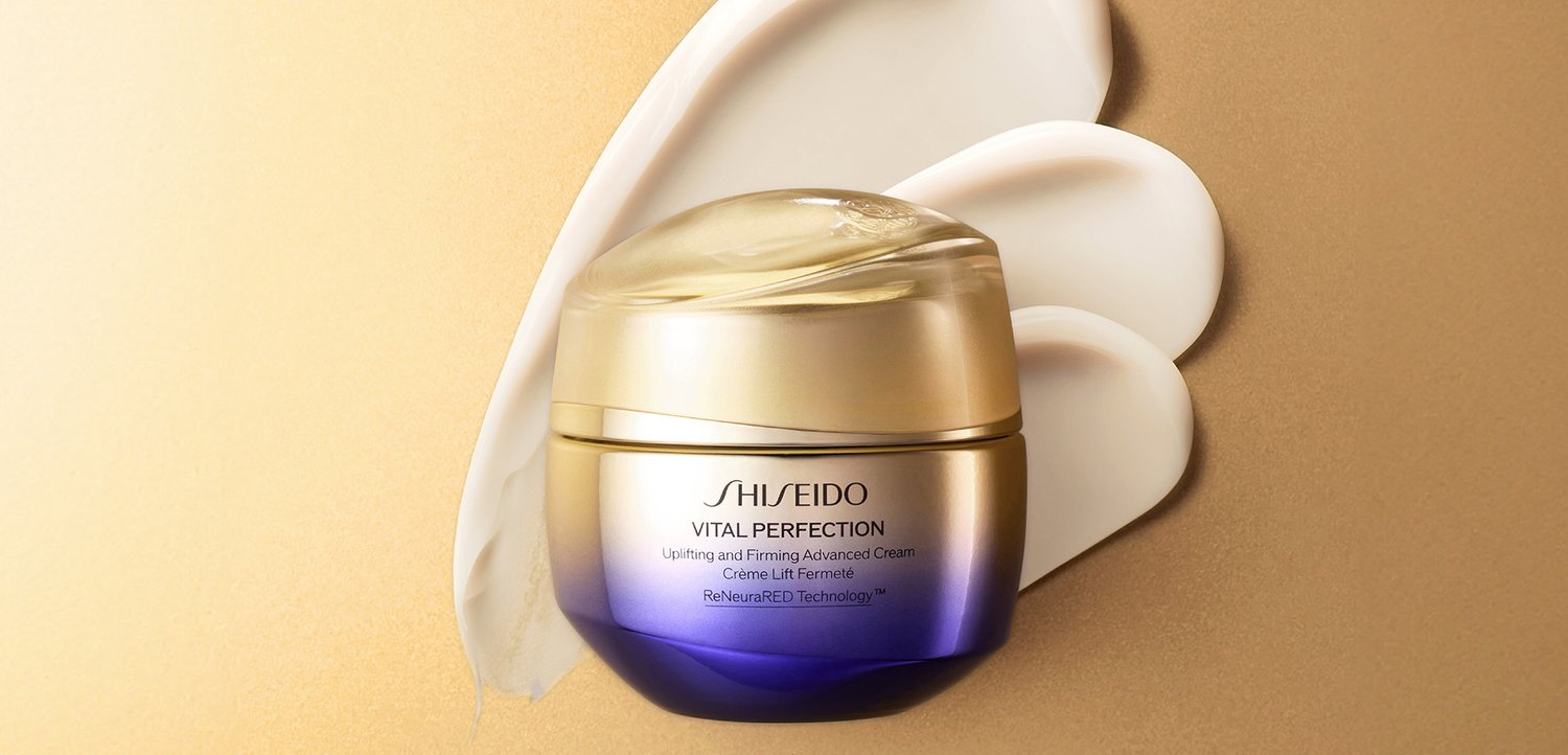 Pot de crème raffermissante Shiseido Vital Perfection avec un emballage doré et violet sur fond beige avec des échantillons de crème en arrière-plan.