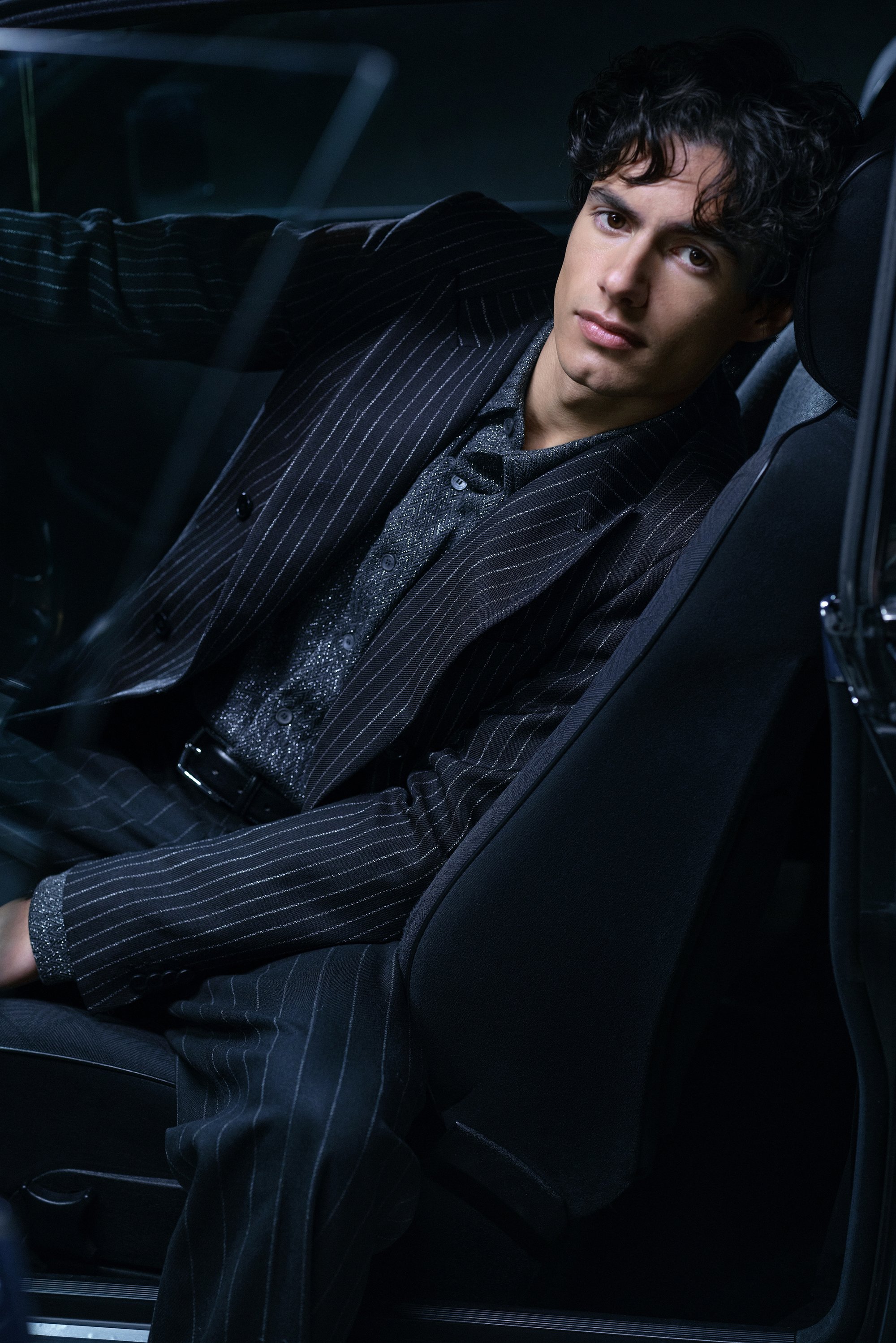 Un joven con cabello rizado vestido con un traje de rayas y una camisa brillante se apoya en el asiento del coche, mirando a la cámara con una iluminación tenue.