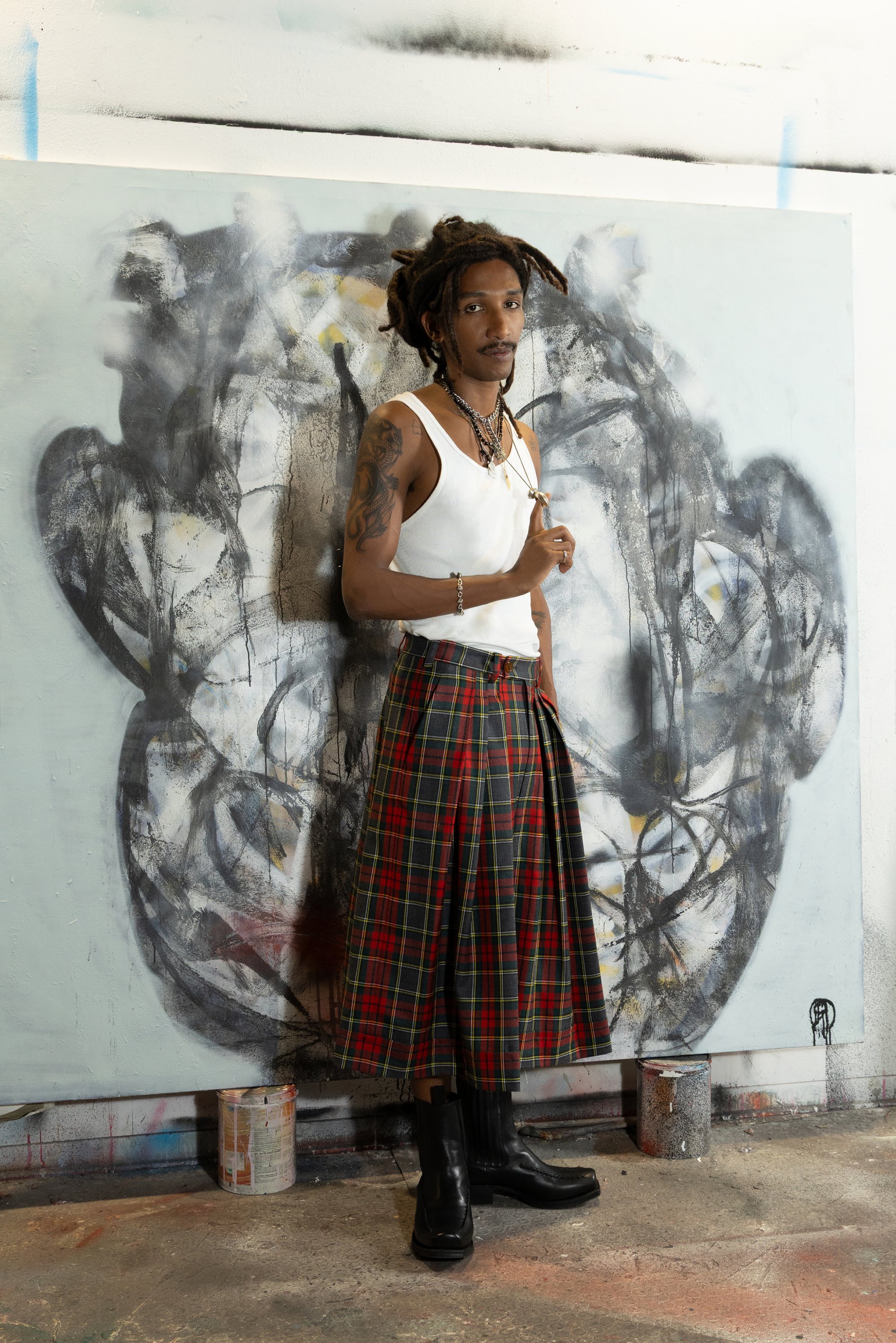 Kilt de cuadros negro y rojo con pliegues, combinado con una camiseta blanca sin mangas y botas de cuero negras. Collares en capas y tatuajes visibles.