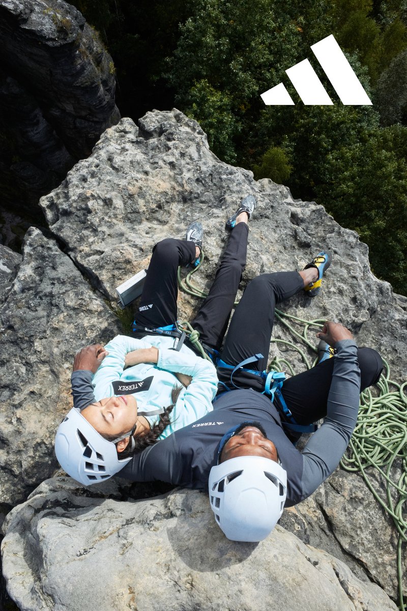 Climbers resting on a rocky ledge, dressed in light blue and dark grey jackets, white helmets, and climbing shoes, with green rope alongside them.