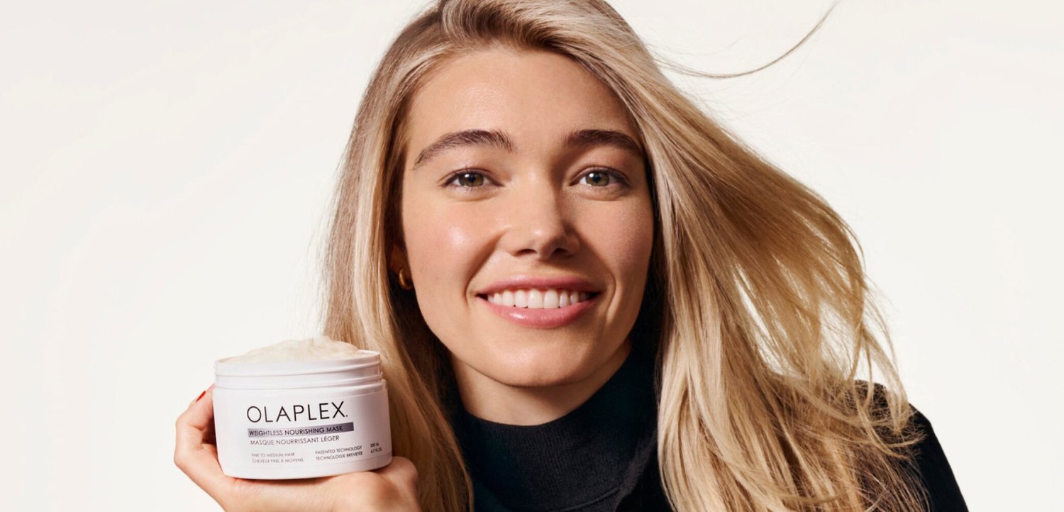 Lachende vrouw met lang blond haar houdt een potje Olaplex haarmasker vast tegen een effen lichte achtergrond.