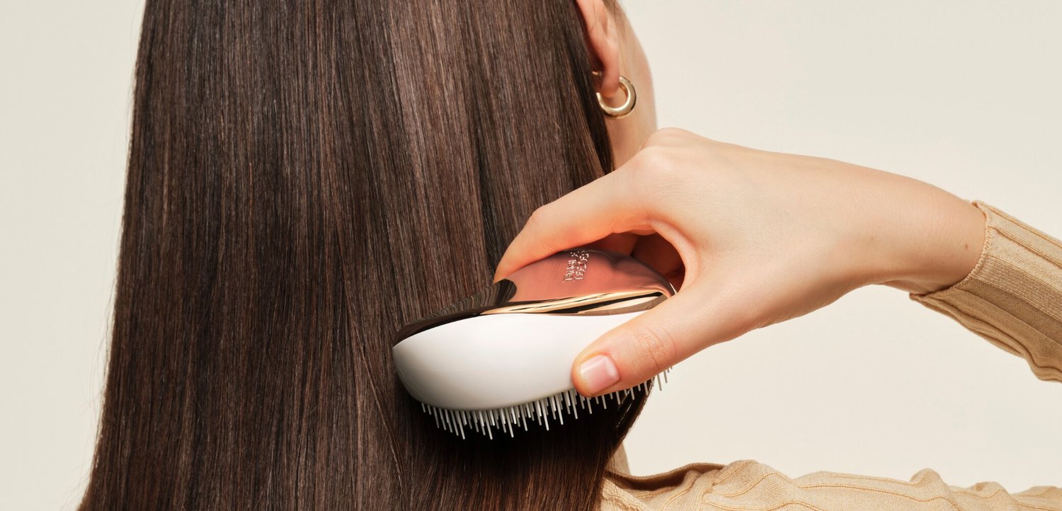 Hand hält eine Haarbürste und kämmt lange, glatte braune Haare vor einem schlichten hellen Hintergrund.