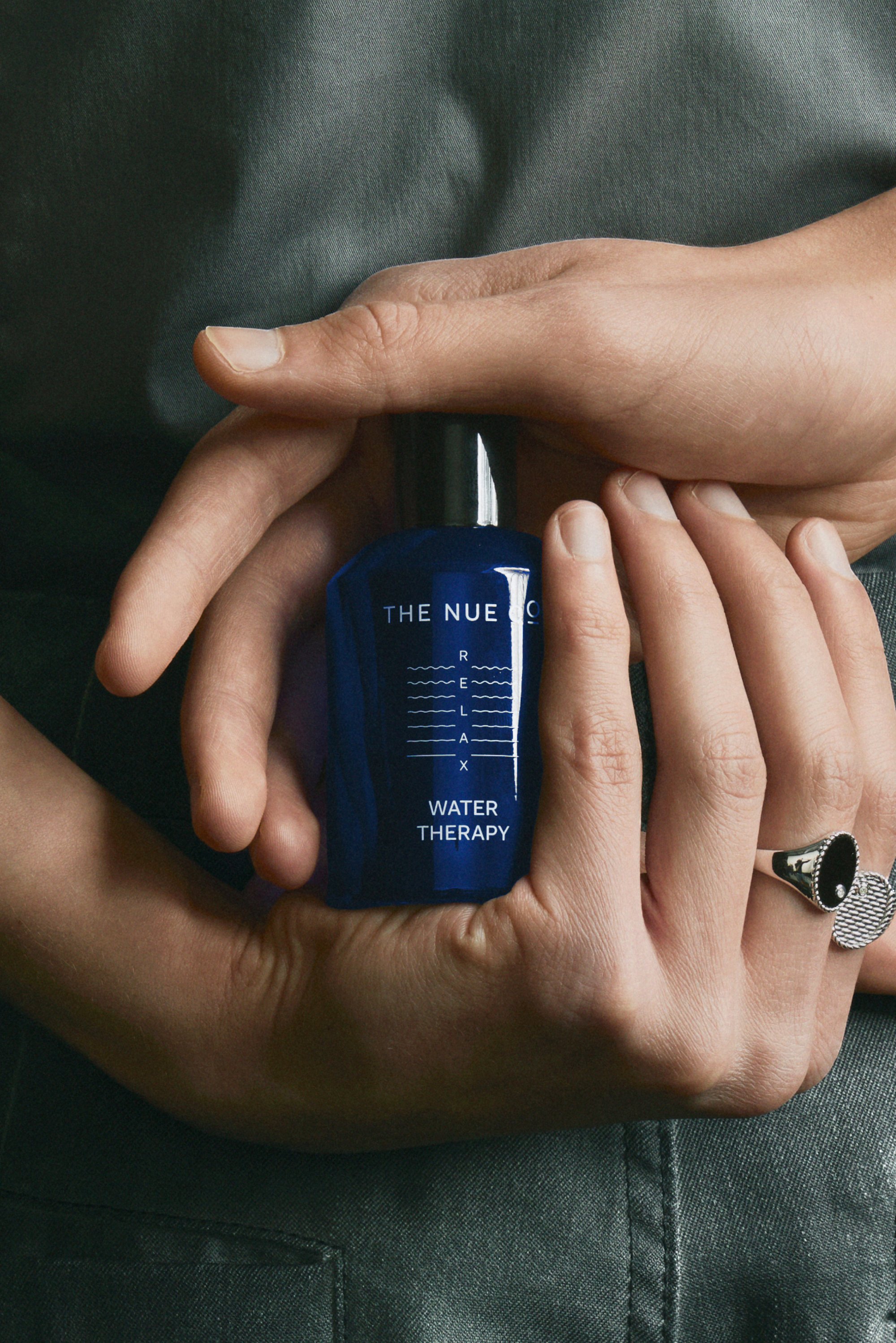 A pair of hands holds a blue bottle labeled "WATER THERAPY" from THE NUE CO, adorned with a silver ring, against a textured grey background.