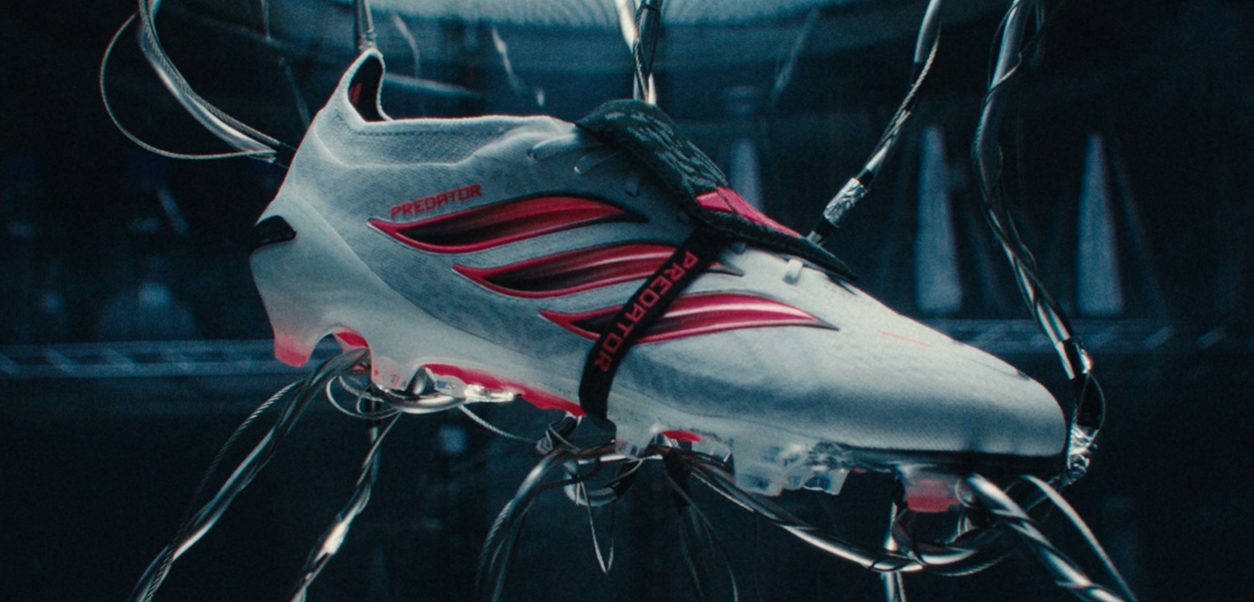 Weißer und roter Adidas Predator Fußballschuh, aufgehängt an schwarzen Drähten in einer dunklen, technologieinspirierten Umgebung.