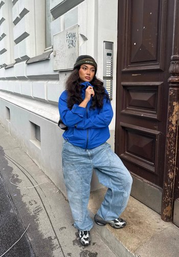 Blå zip-up jacka med hög krage, överdimensionerade ljusblå jeans och svart-vita mönstrade sneakers. Stående mot en texturerad vägg.