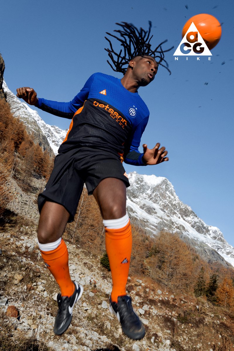 Athlete wearing orange and black kit jumps mid-air outdoors with snowy mountains and autumn trees in the background under clear blue sky.