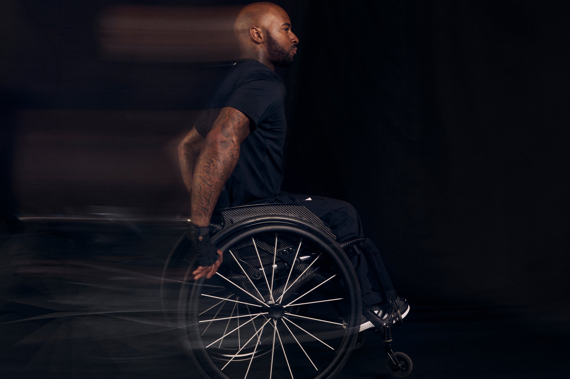 A strong athlete with tattoos propels a wheelchair forward, focused, against a dark background, conveying determination and motion.
