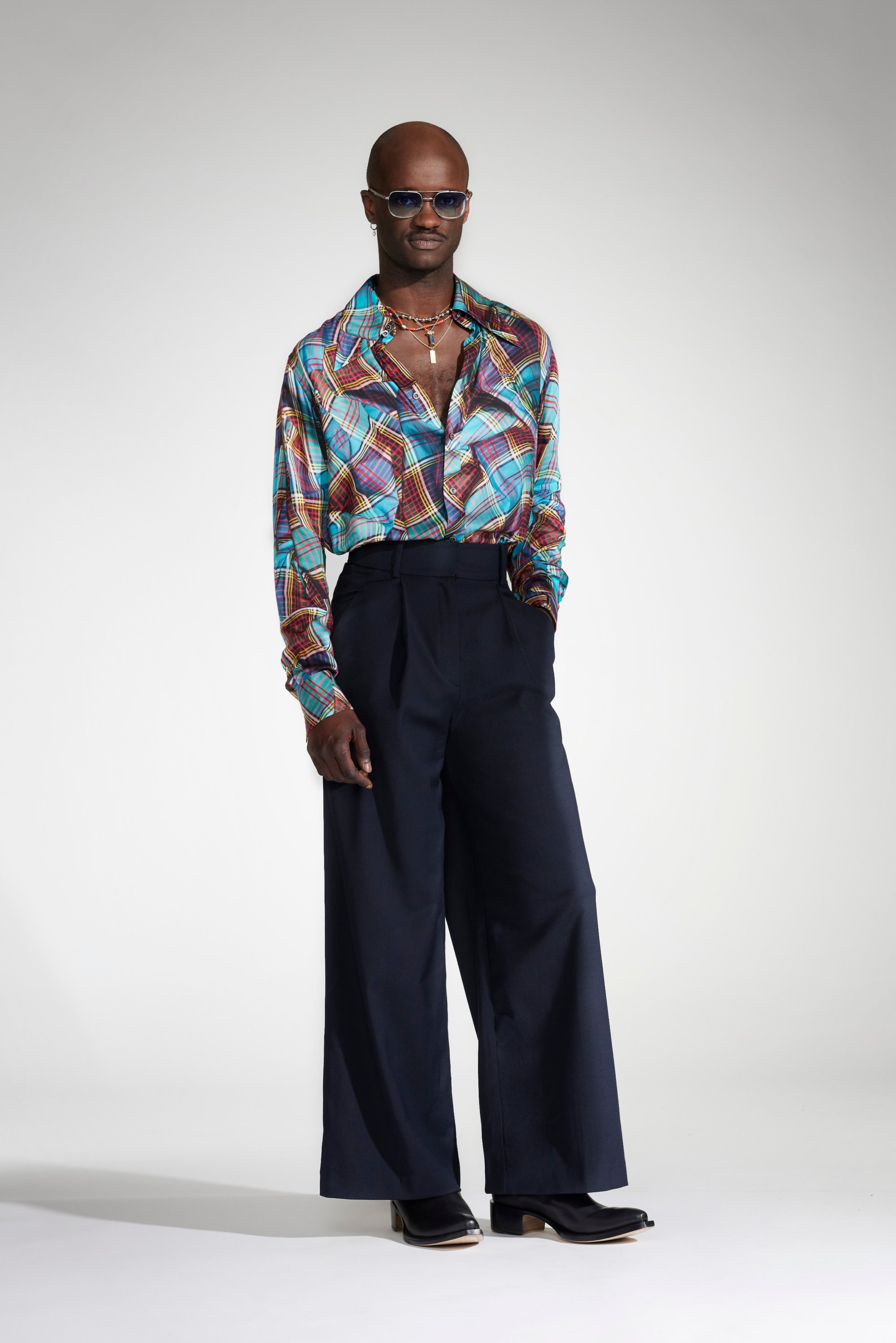 A person dressed in a vibrant patterned shirt and wide-leg black trousers, standing with confidence, arms relaxed by their sides, wearing fashionable sunglasses.