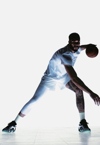 Un joueur de basket-ball en uniforme blanc effectue un dribble dynamique, mettant en valeur des tatouages sur ses jambes contre un fond blanc éclatant.