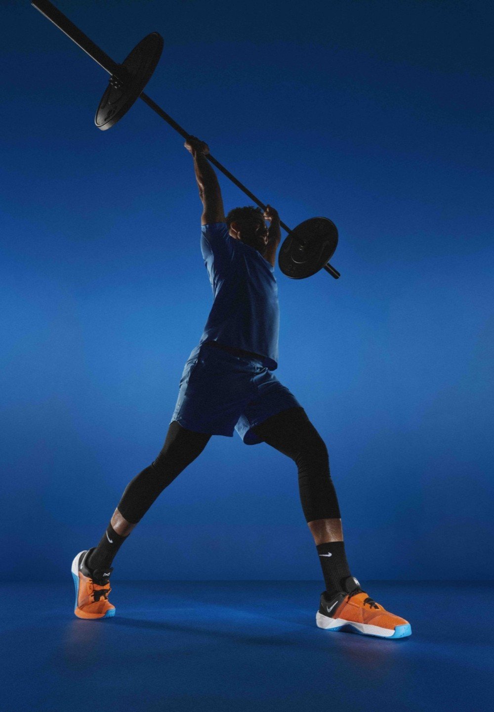 Halteres noires levées au-dessus de la tête par une personne en chemise bleue, short et leggings noirs, portant des chaussures de sport orange et noires. Fond bleu.