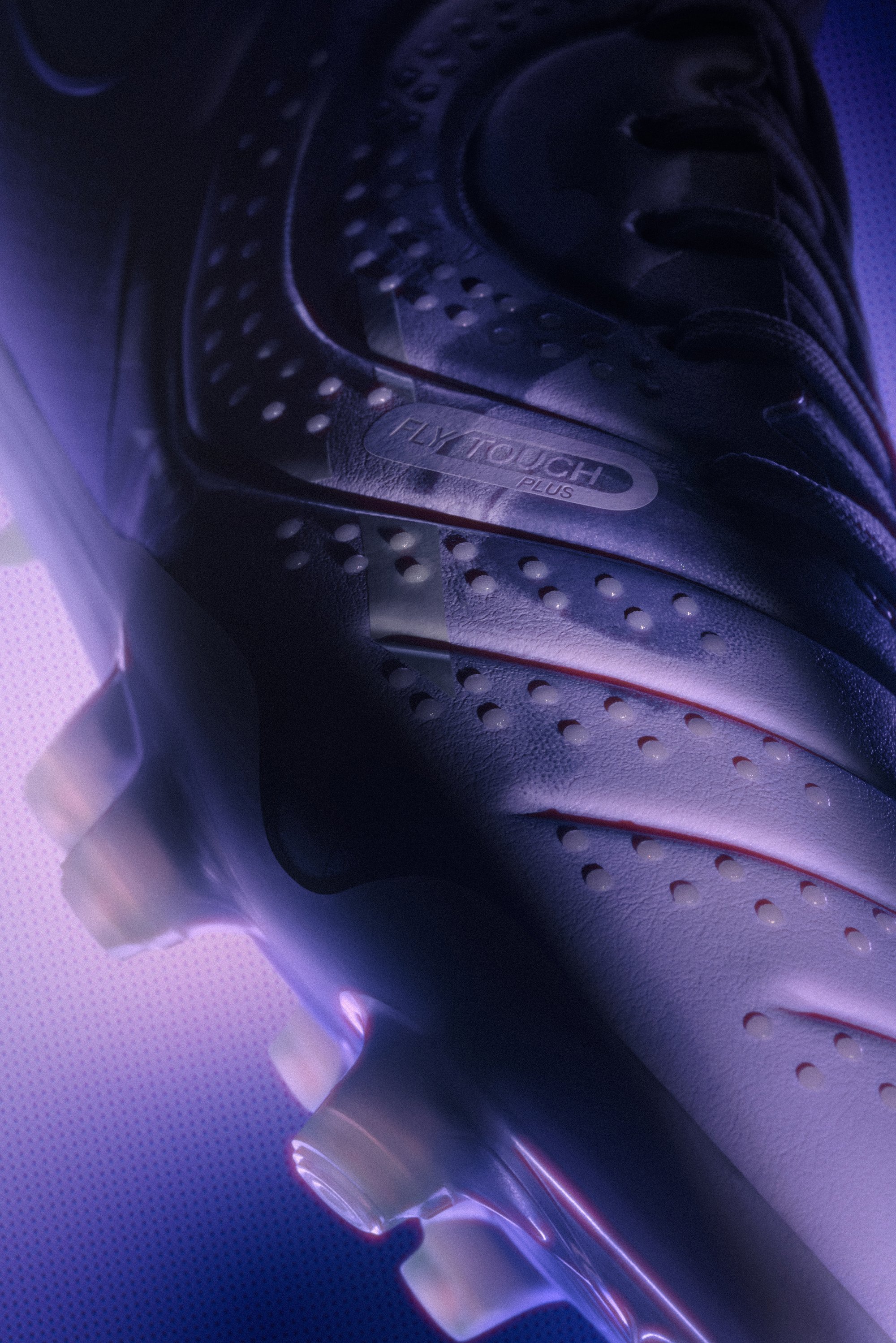 Acercamiento de una elegante bota de fútbol negra con la marca "FLYTOUCH PLUS", contra un fondo iluminado en púrpura.