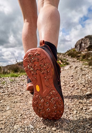 Person walking on rocky trail wearing hiking shoes with mud on the sole, visible calf muscles, and partly cloudy sky.