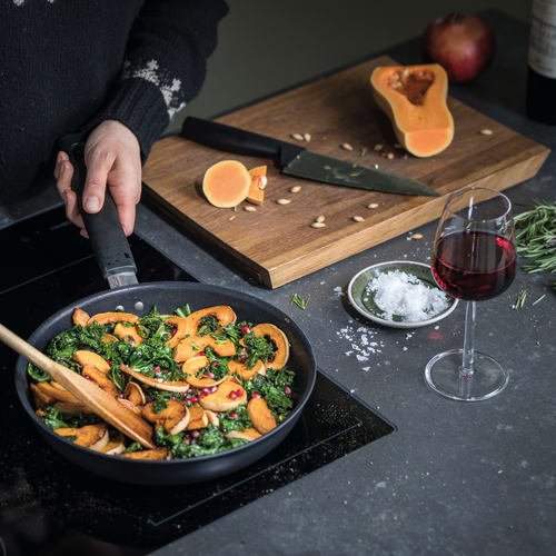 Czarna patelnia z podsmażonym zielonym jarmużem i plasterkami pomarańczowego dyni, udekorowana czerwoną papryczką. W tle szklanka czerwonego wina oraz drewniana deska do krojenia.