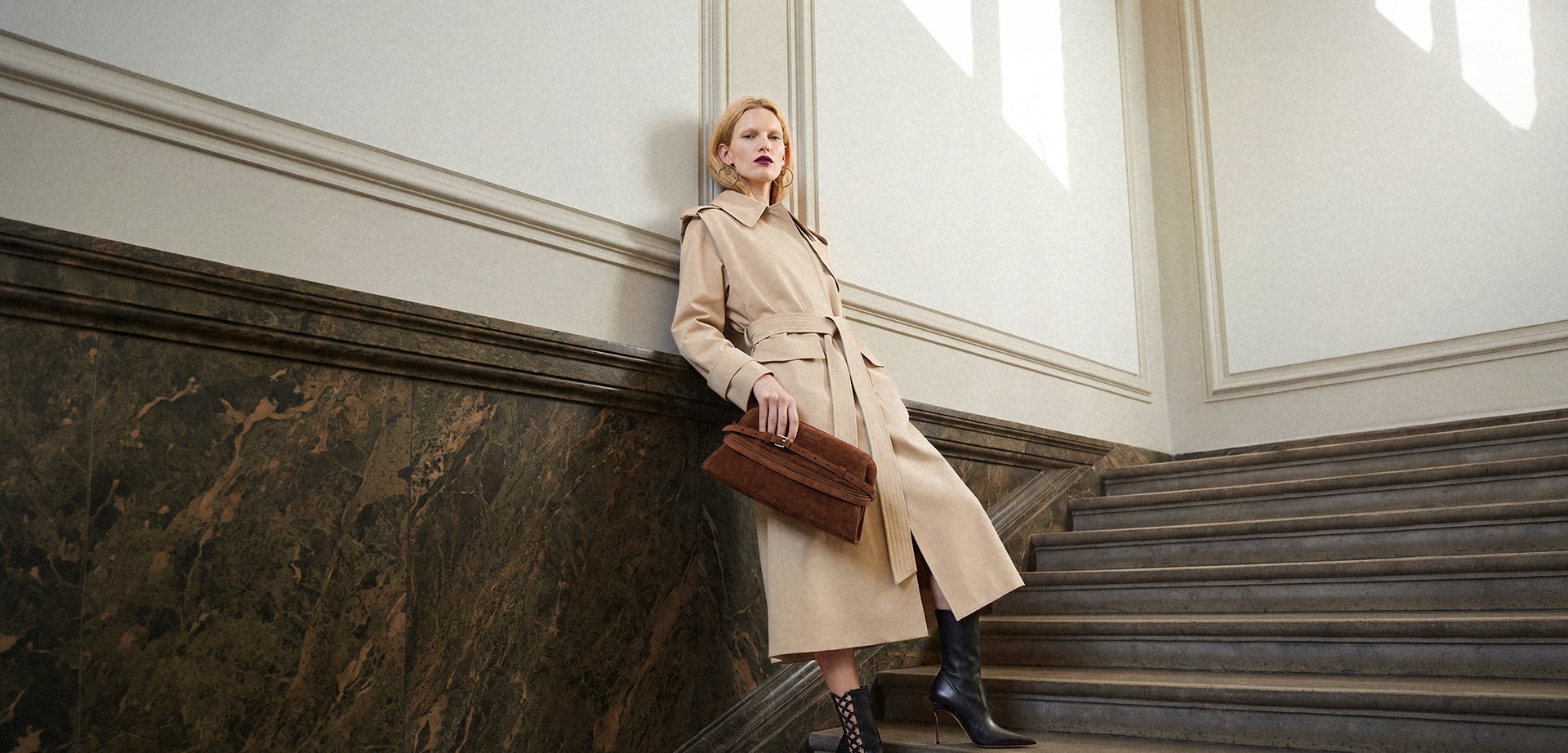 Femme en trench beige et bottes noires s'appuie contre un mur en marbre en tenant une pochette marron près d'un escalier dans un espace bien éclairé.