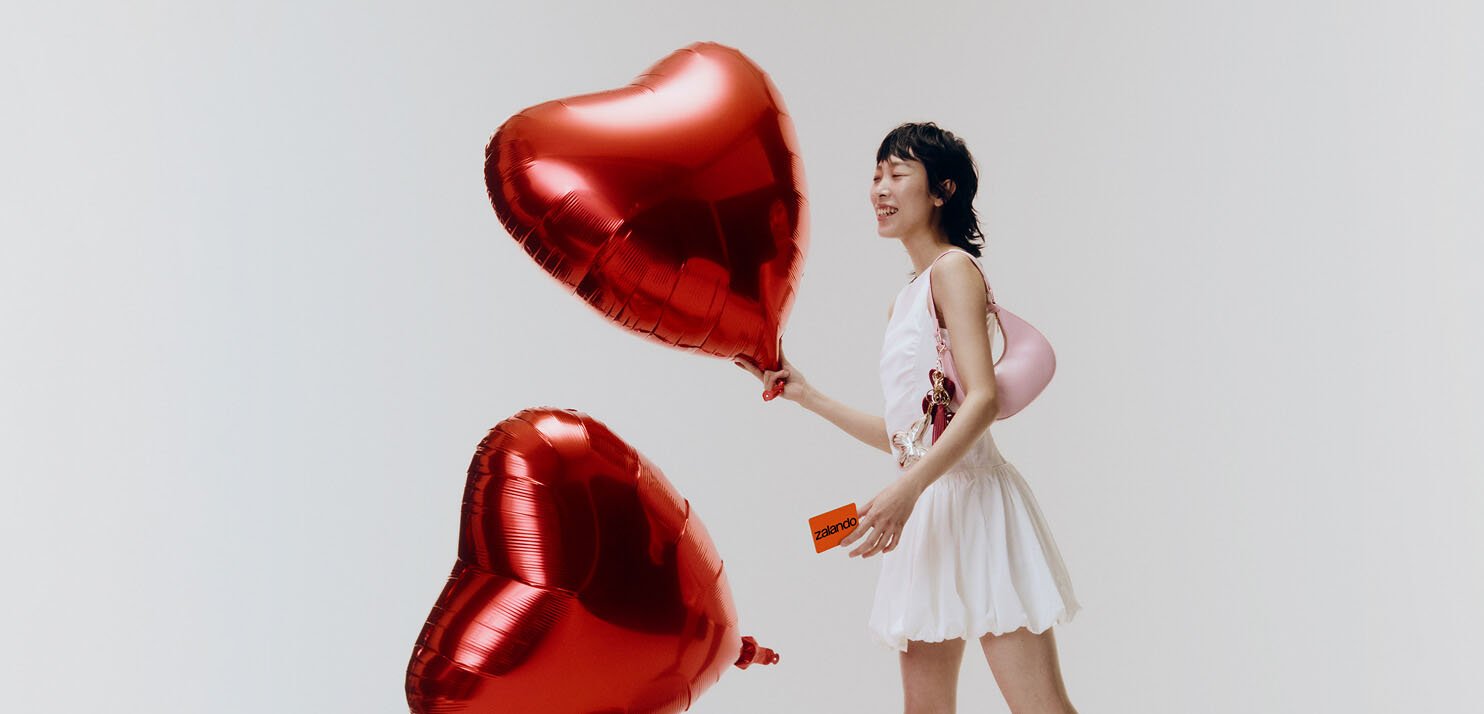 Mlada ženska v beli obleki drži dve rdeči balončki v obliki srca in orančno kartico Zalando, nasmejana na belem ozadju.
