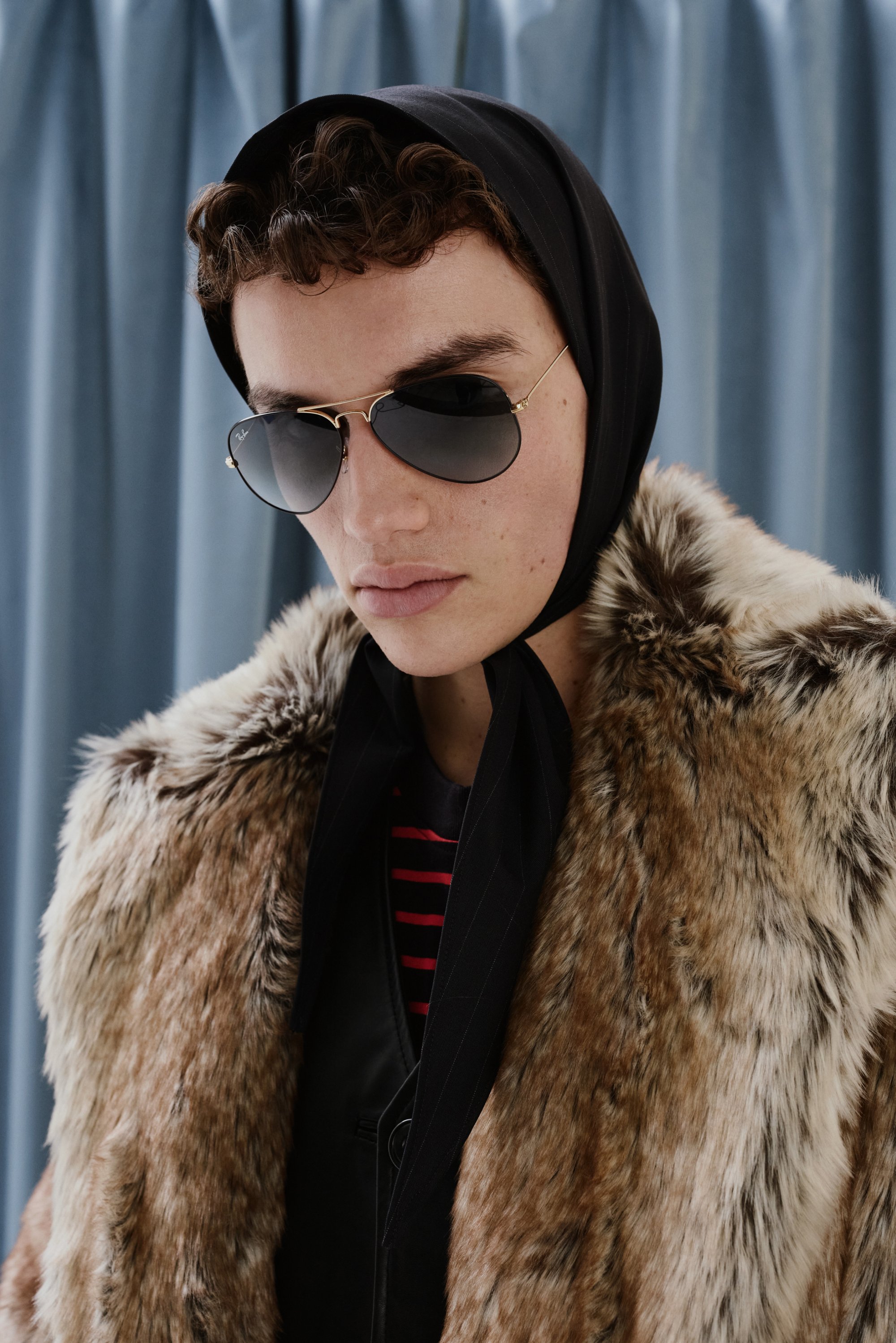 A person with curly hair wears sunglasses and a black scarf over their head, styled in a fur coat against a blue curtain backdrop.
