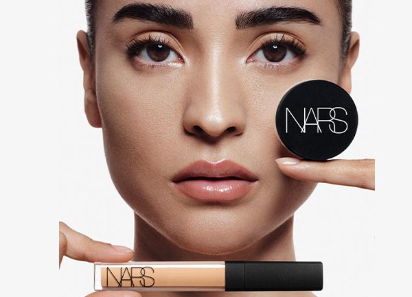 Model holds a concealer and pressed powder compact labeled "NARS," showcasing a natural makeup look with flawless skin.