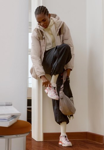 Woman with braided hair tying white and pink trainers indoors, wearing a beige jacket, black trousers, and carrying a grey crossbody bag.