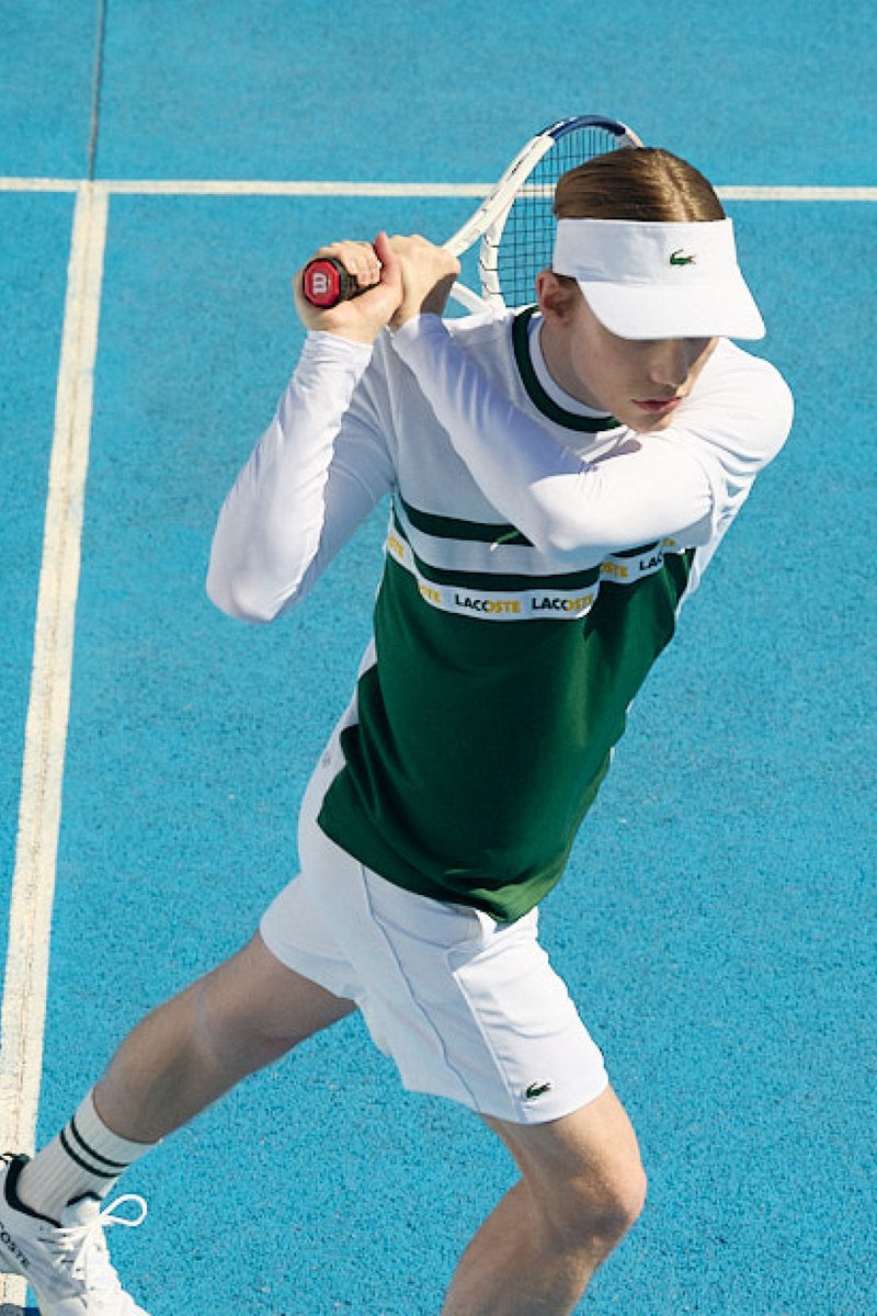 Una giovane giocatrice di tennis in un completo verde e bianco si prepara a servire, mentre un giocatore maschio in blu attende pronto su un campo blu.