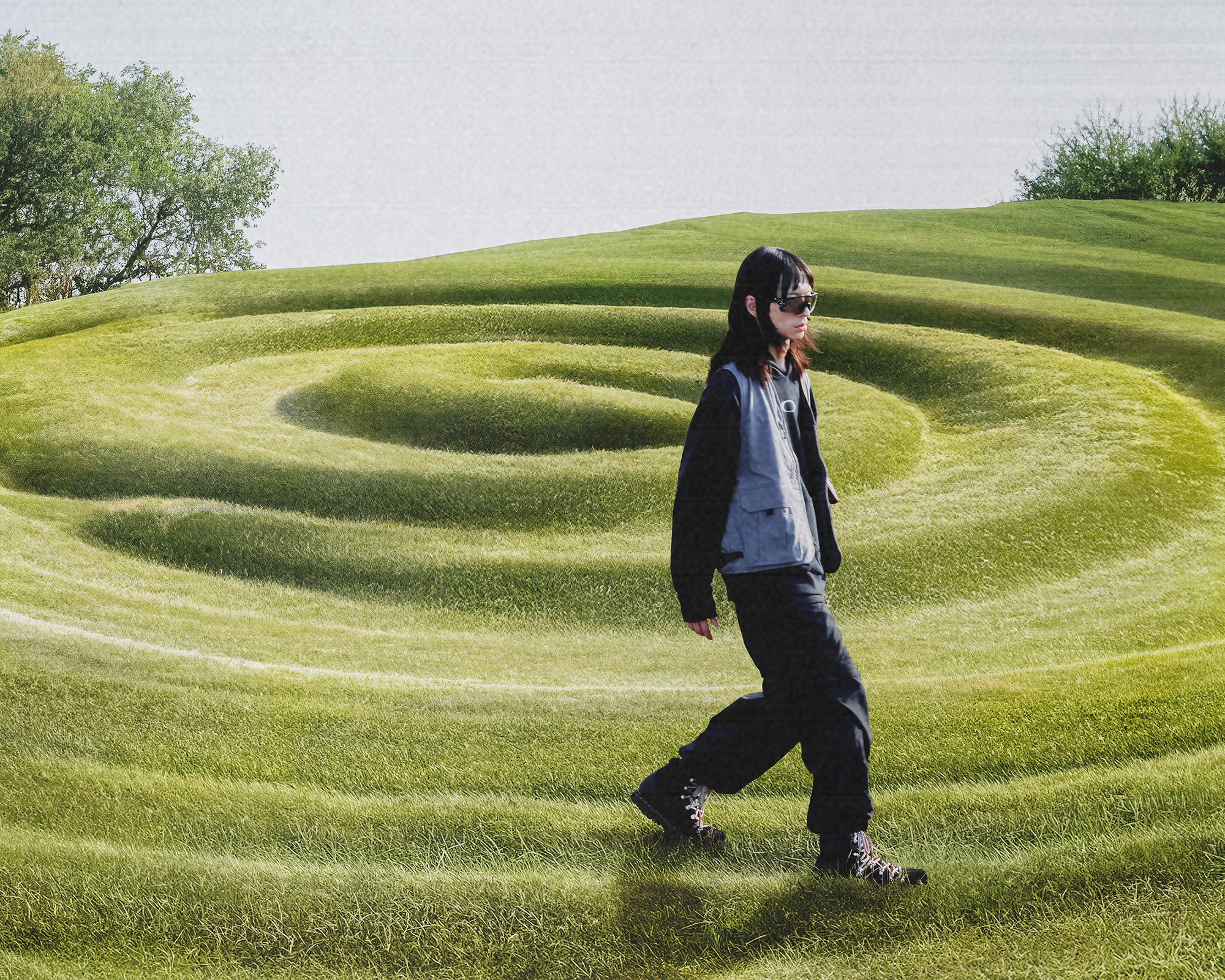 A person in sunglasses walks along a spiral-patterned grassy landscape under a bright sky, with trees in the background.