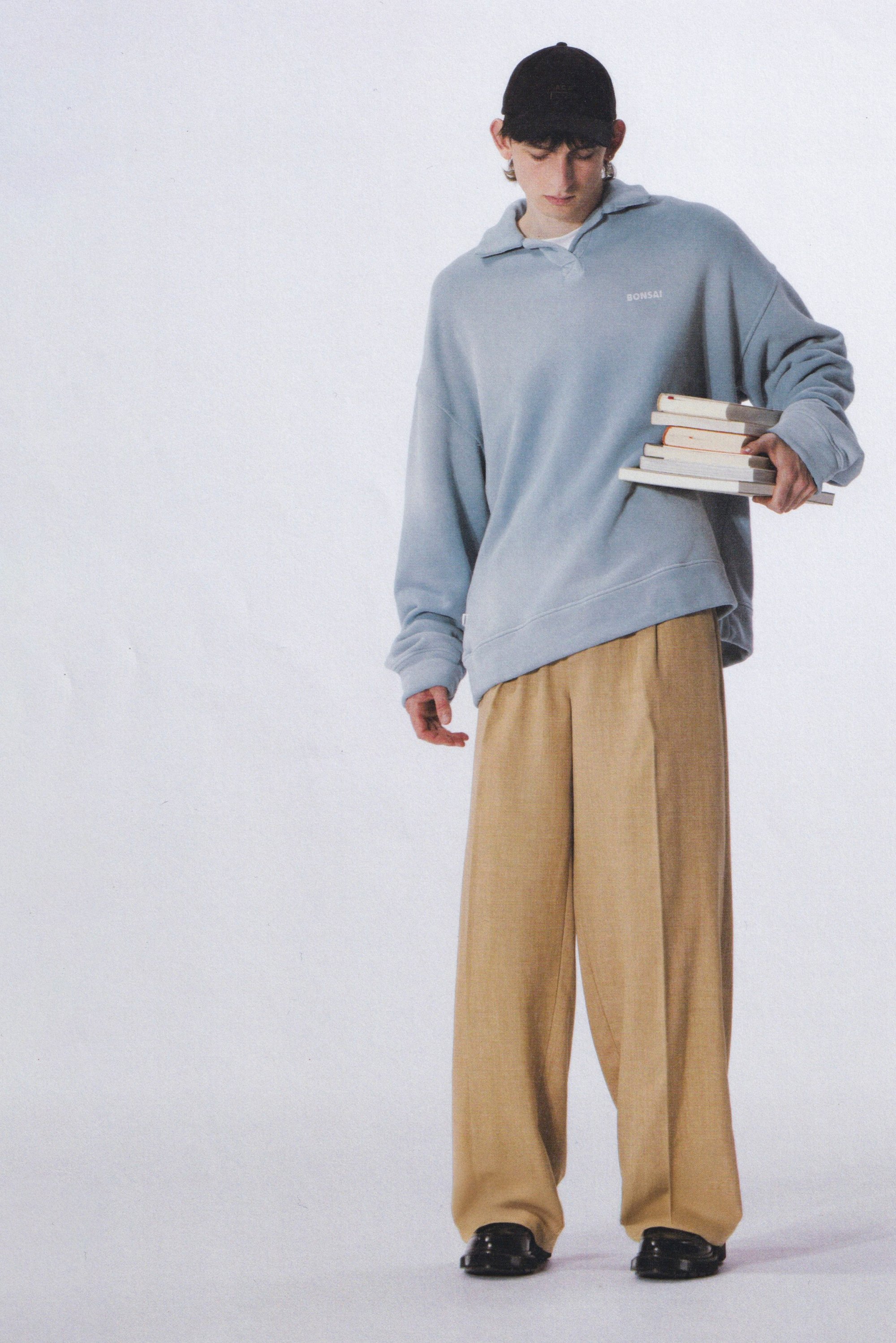Lichtblauwe oversized hoodie met een kraag en logo, gecombineerd met beige wijde broekspijpen. Houdt in één hand verschillende opgestapelde boeken vast.