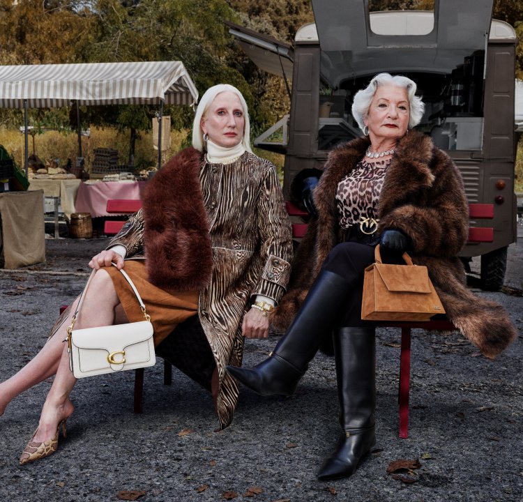 Two women are seated on benches. One is wearing a patterned coat and has a white handbag, while the other is dressed in a fur coat and carrying a brown handbag.
