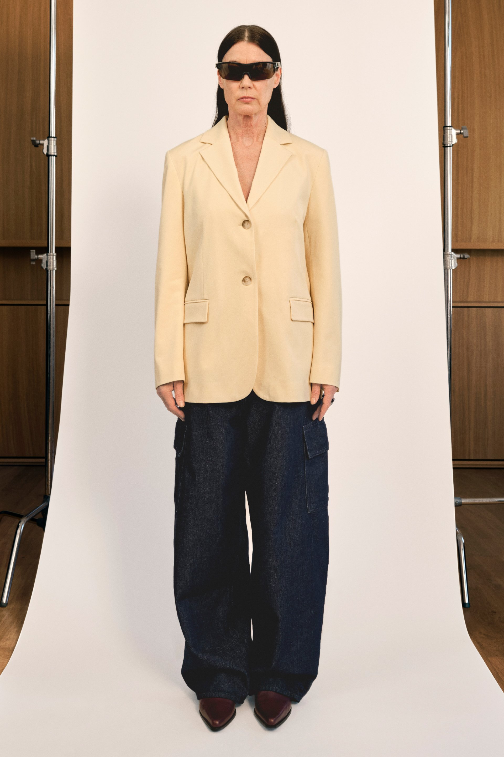 Una mujer con cabello largo y oscuro lleva puesta una chaqueta amarilla, pantalones cargo oscuros y gafas de sol, de pie contra un fondo liso en un estudio.
