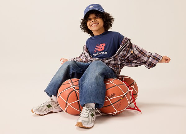 Blå New Balance-keps, marinblå T-shirt med logotyp, rutigt oversized skjorta, denimjeans och vita/gröna sneakers; sitter på två basketbollar.