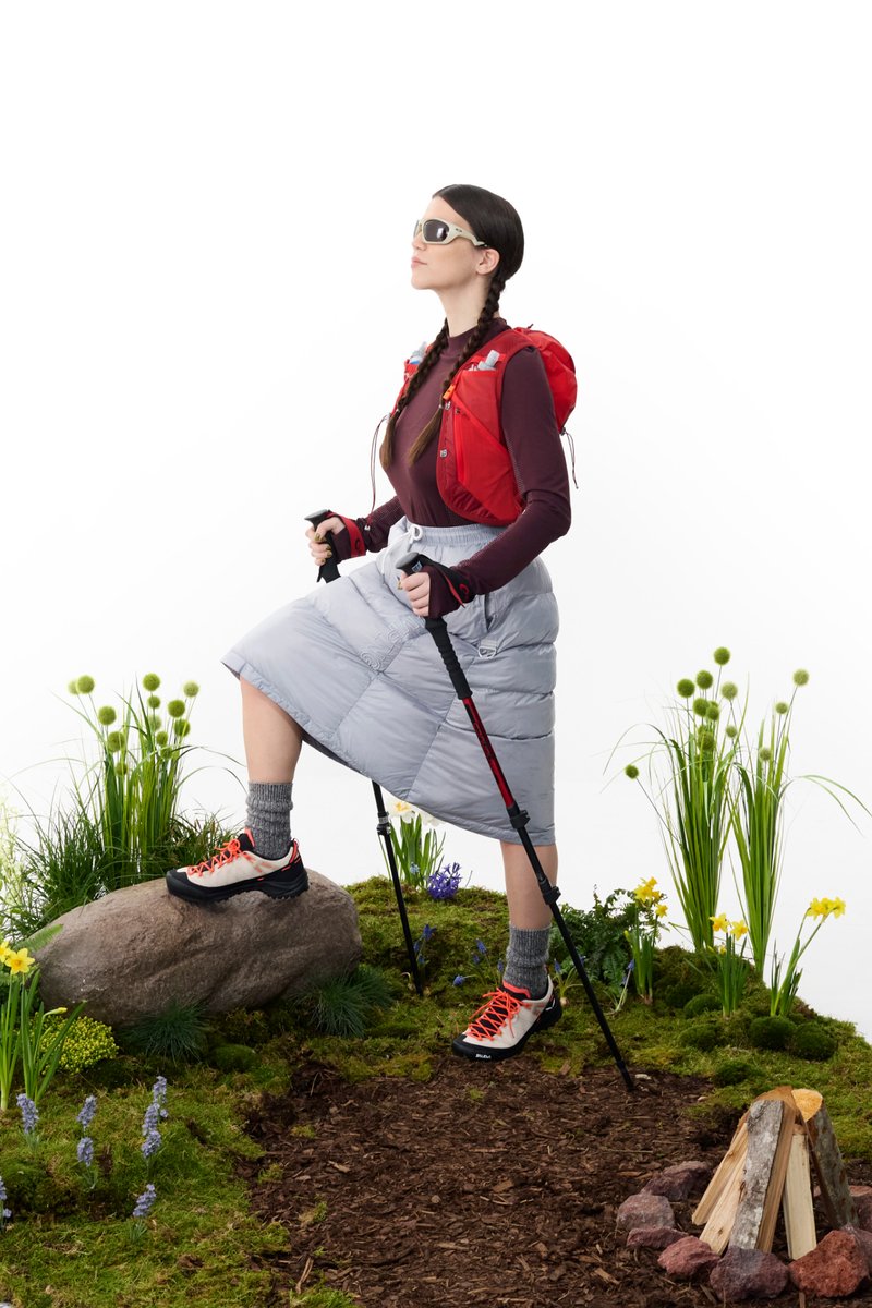 Una donna con occhiali da sole beve da una tazza da viaggio mentre una persona con un cappotto verde osserva la natura attraverso dei binocoli. Intorno a loro ci sono vegetazione e un'area attrezzata con un falò.