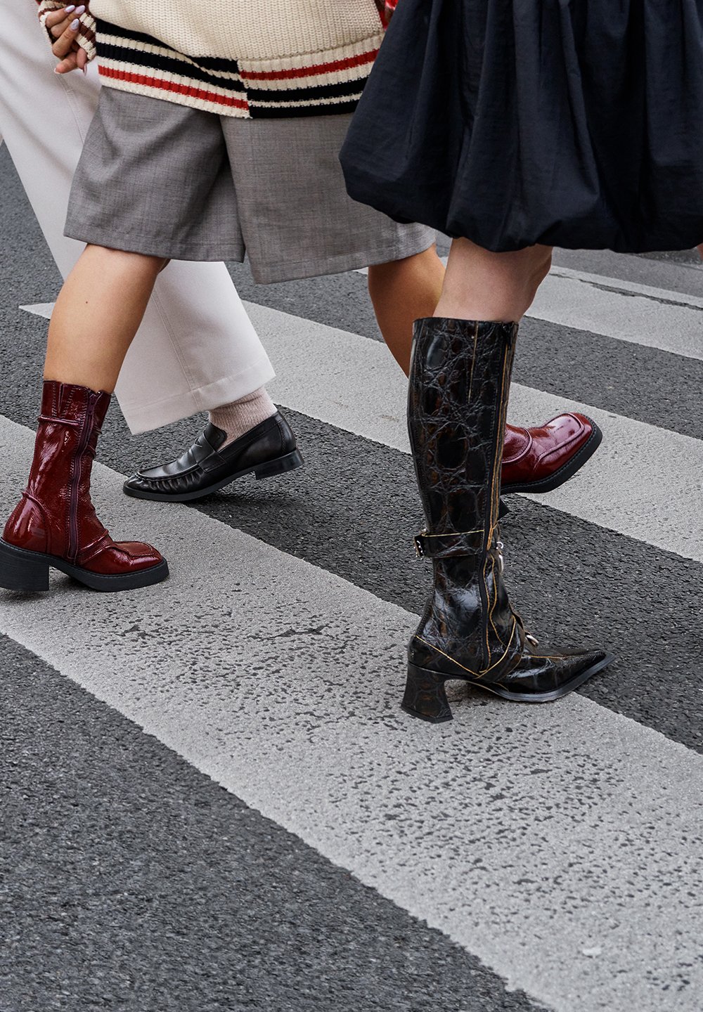 Three pairs of footwear on a pedestrian crossing: a shiny red ankle boot, a black leather flat shoe, and a textured black knee-high boot with a block heel.