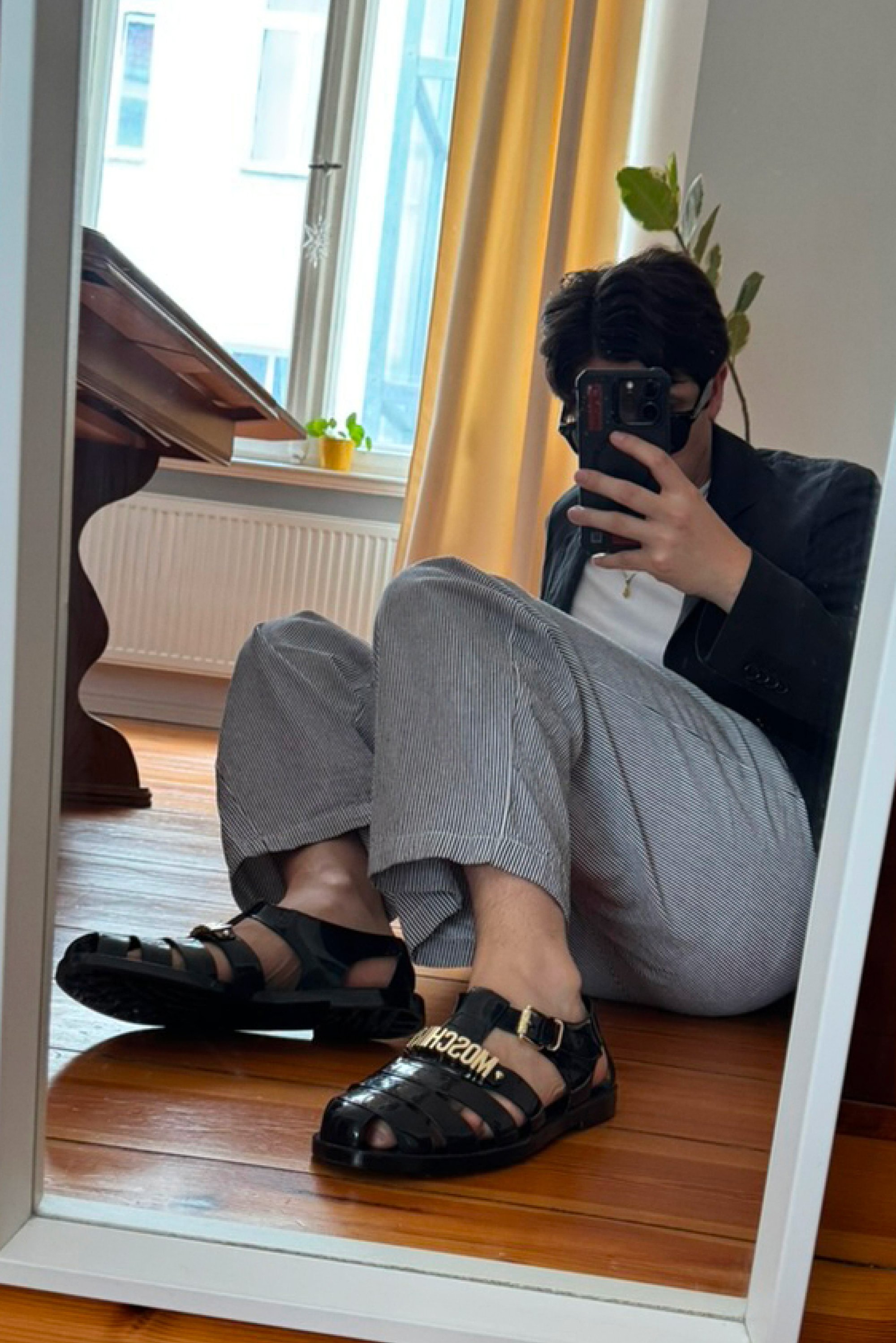 Sandali neri in plastica con un design a strisce, caratterizzati da scritte dorate e un dettaglio con fibbia. Pantaloni a righe e uno specchio riflettente presentano le calzature.