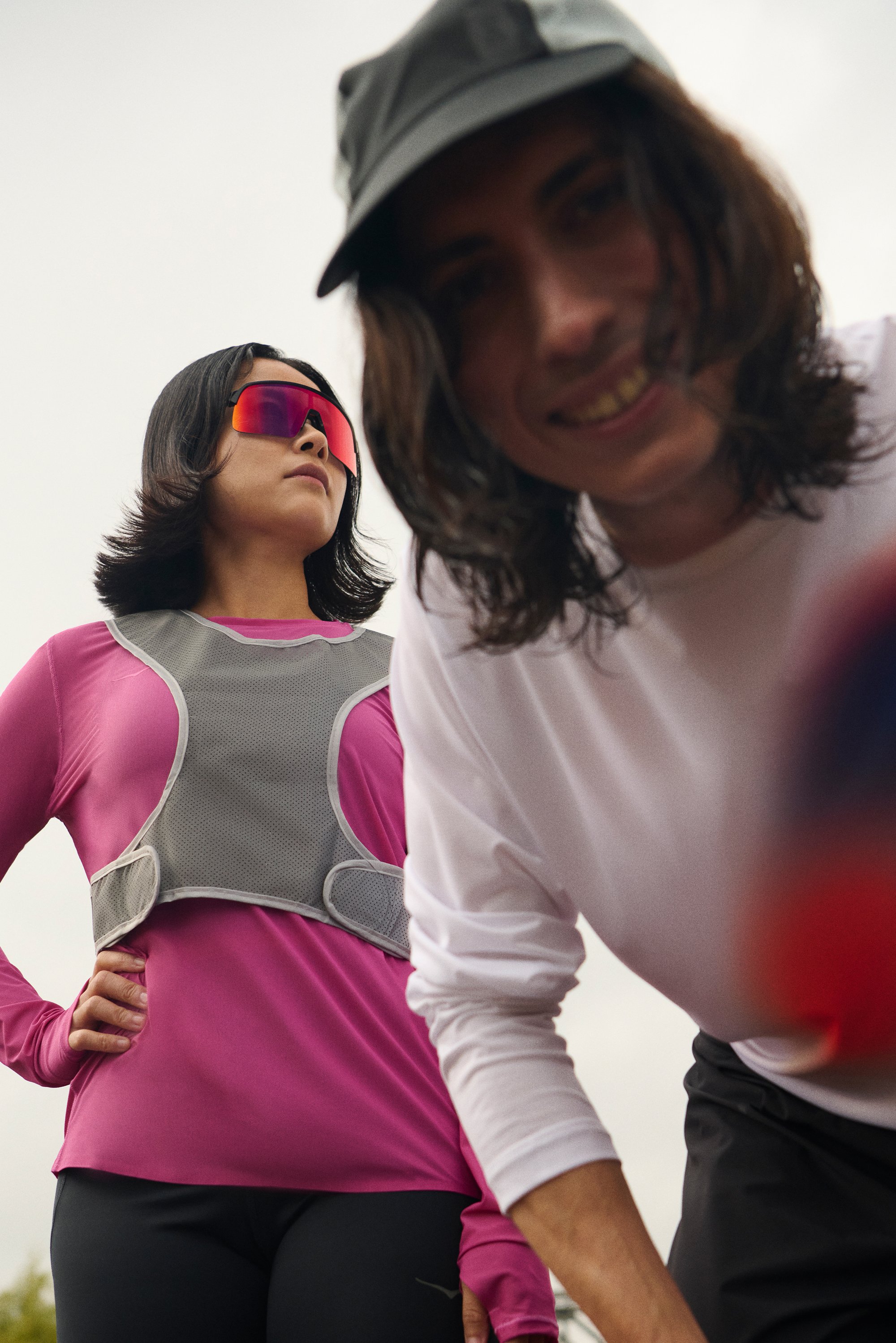 A woman in a pink long-sleeve shirt and sunglasses stands confidently, while a smiling man in a white shirt leans towards the camera.