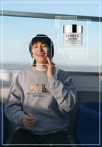 Clinique Even Better lysende fuktighetskrem i en hvit krukke med s ølvlokk. Person har på seg en grå genser med broderte hunder.