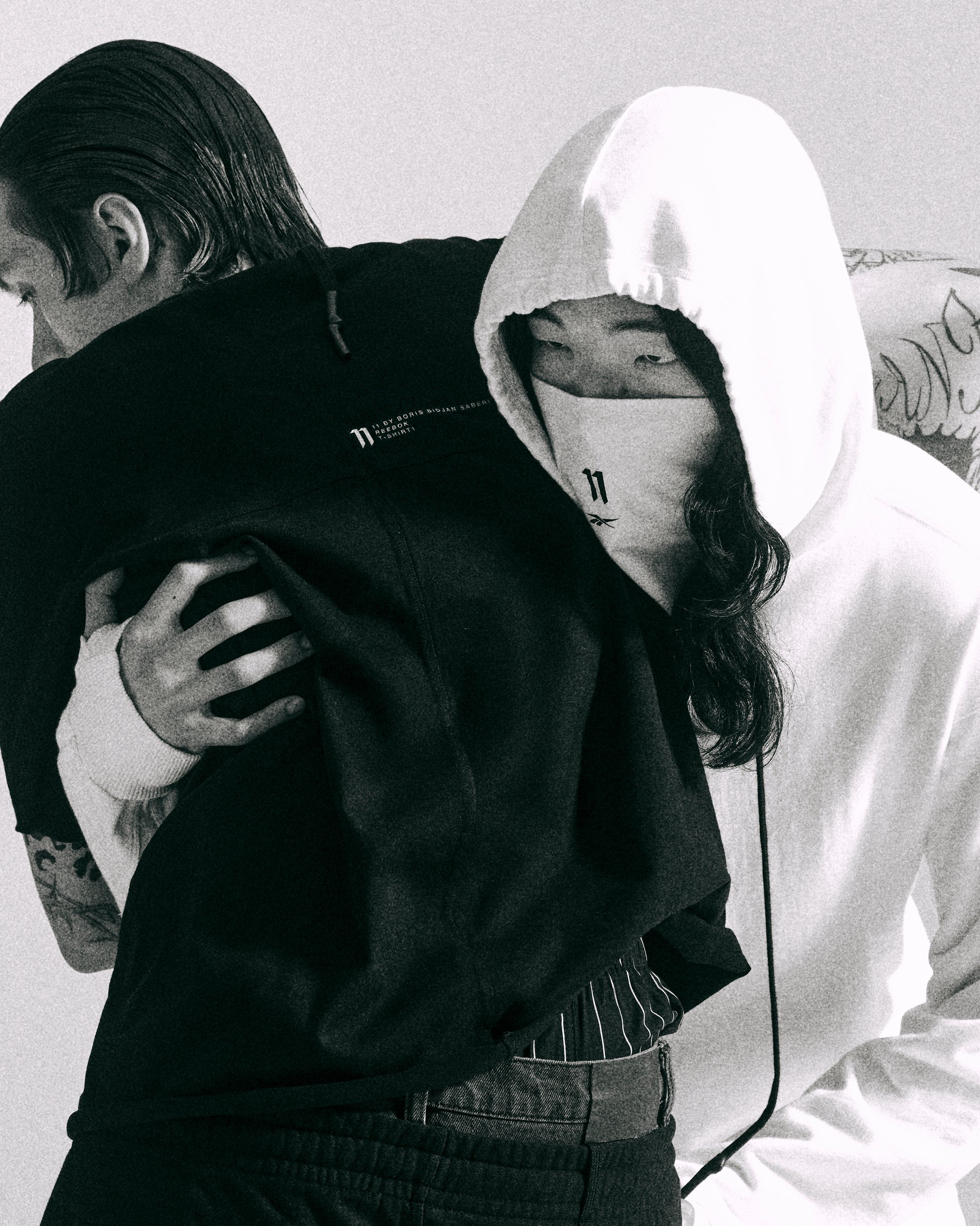 Dos personas se abrazan, una vistiendo una sudadera negra y la otra con una sudadera blanca y una máscara, creando una atmósfera íntima y melancólica.