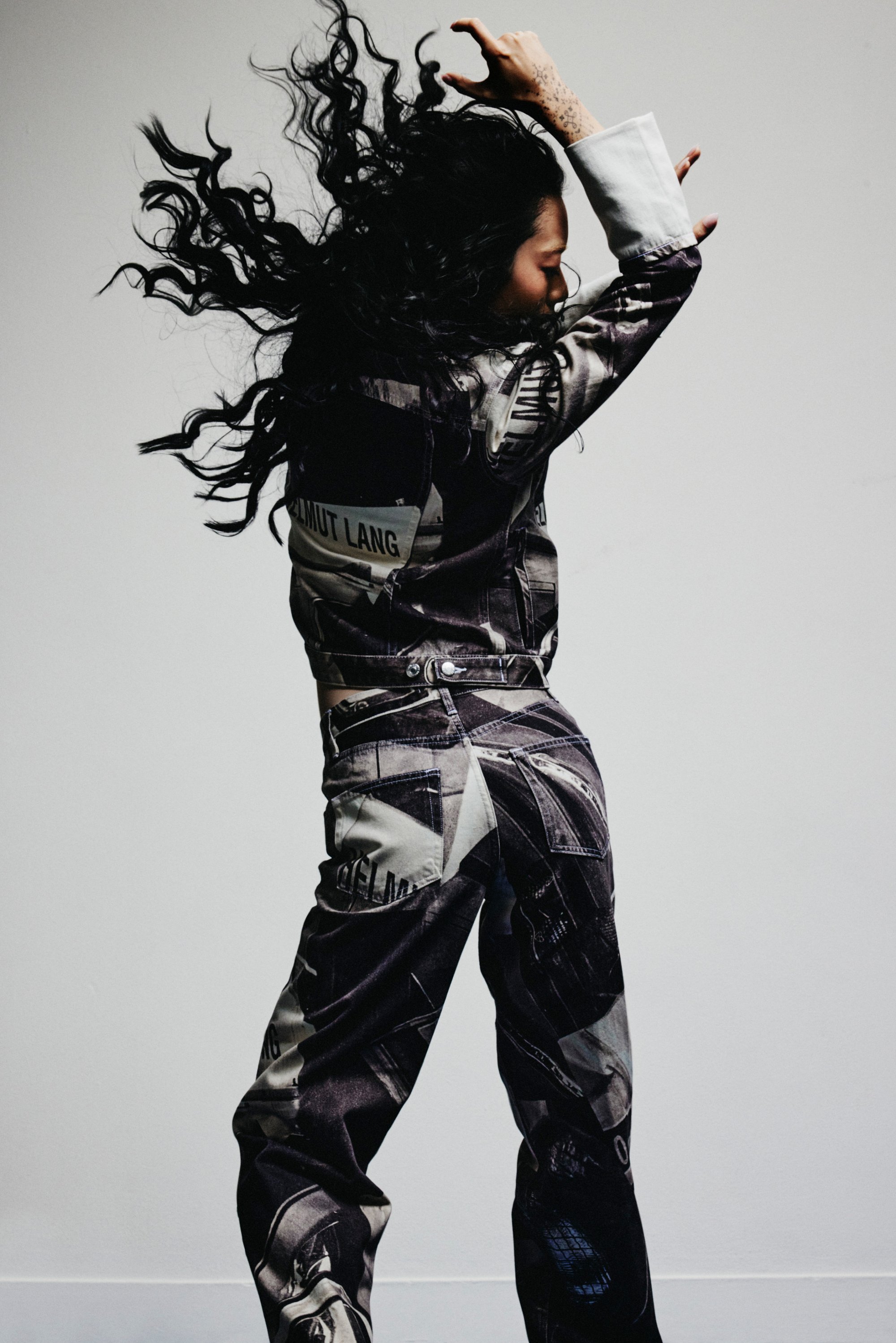 Una persona con cabello largo y ondulado baila, vistiendo un conjunto a rayas en blanco y negro. El fondo es sencillo, destacando el movimiento dinámico.