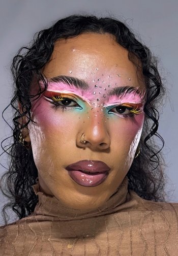 Person mit lockigem schwarzen Haar trägt mutiges pastellfarbenes Augen-Make-up mit gelben Wimpern, pink gefiederten Augenbrauen, Strasssteinen auf der Stirn und glänzenden dunklen Lippen.